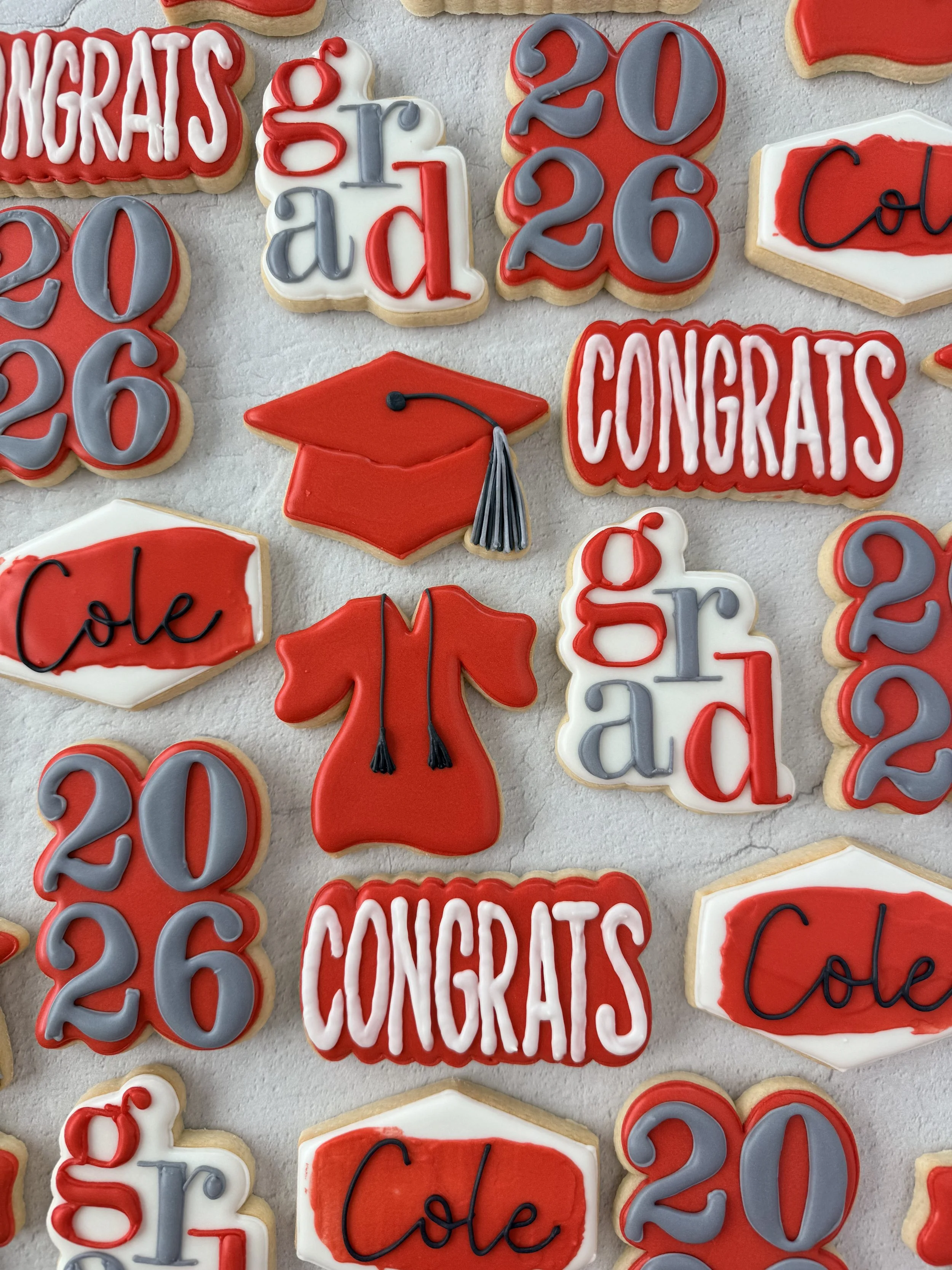 Cookies decorated with red, gray, white, and black icing spelling out 'CONGRATS,' '20,' 'grad,' 'class,' '2023,' and 'yole.' one cookie has a red graduation cap with a gray tassel.