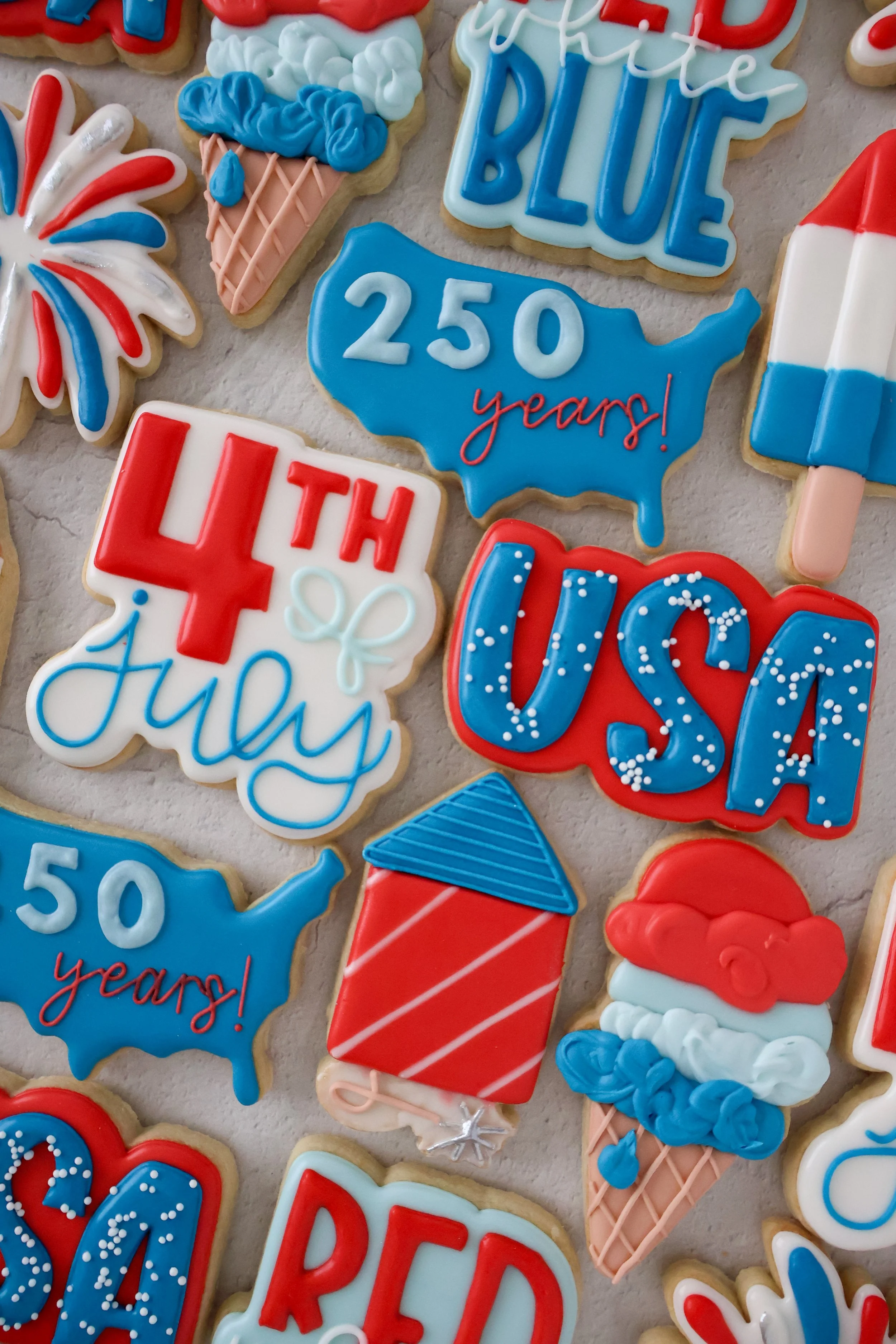 Celebratory cookies with patriotic red, white, and blue colors, including ice cream cones, fireworks, the number 250, a Texas shape, and various other festive designs.