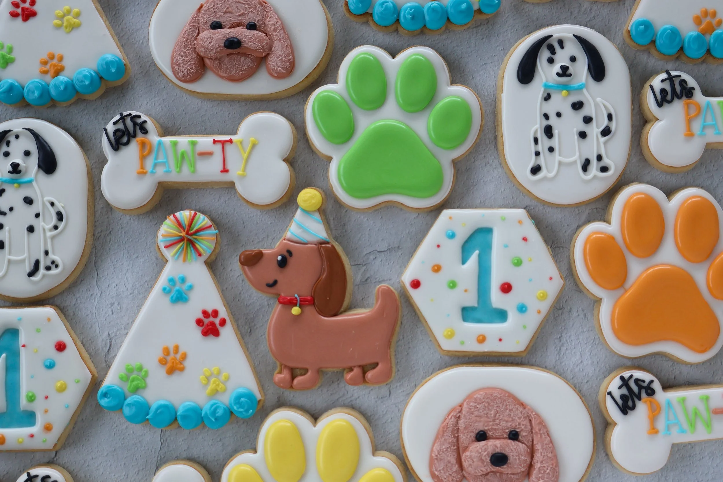 Collection of decorated cookies with a dog theme, including cookie with a dog face, green paw print, and birthday-themed cookies with colorful decorations and lettering.