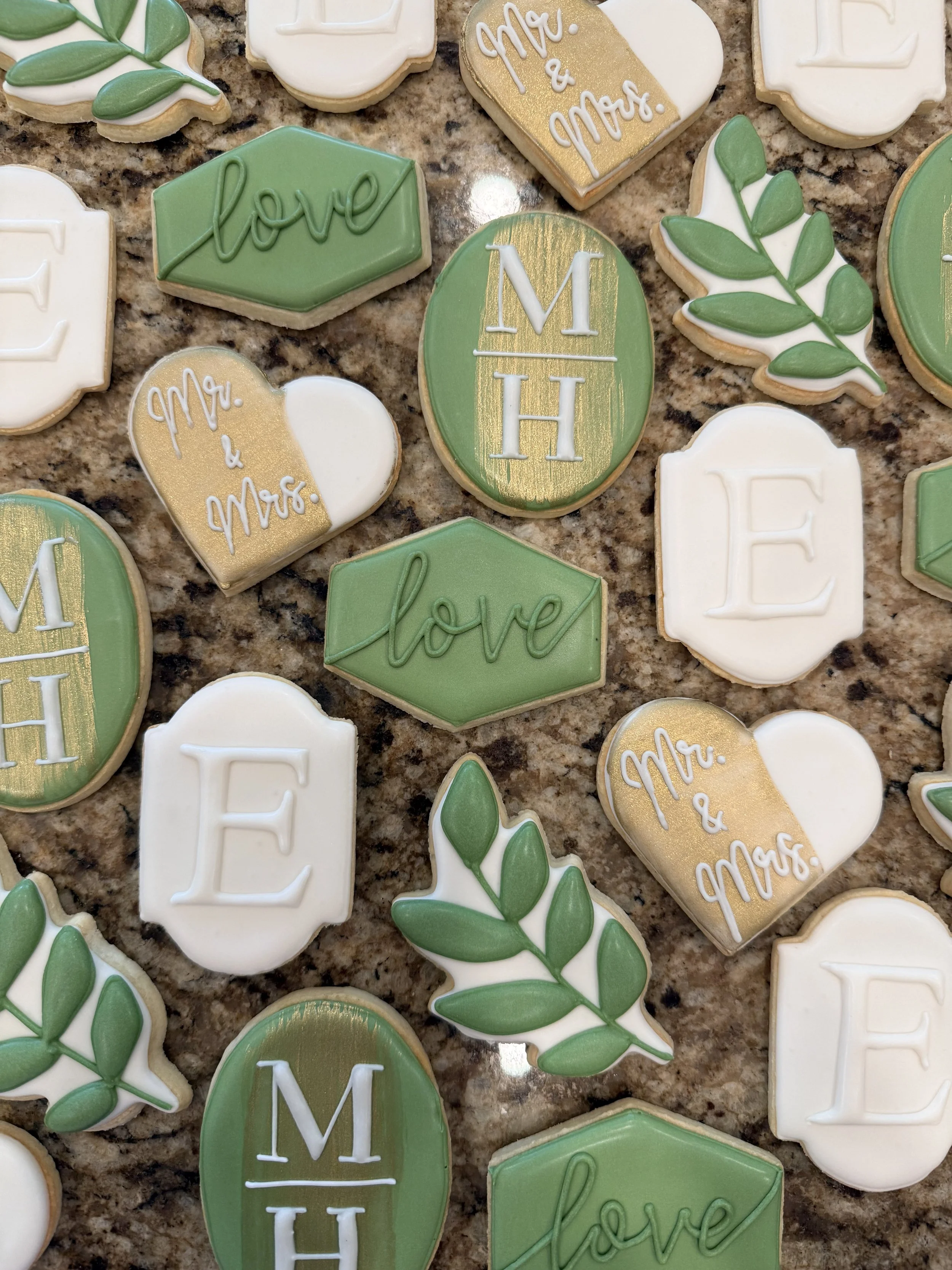 Decorative cookies with various messages and designs, including hearts, leaves, and lettered initials for a wedding or romantic celebration.