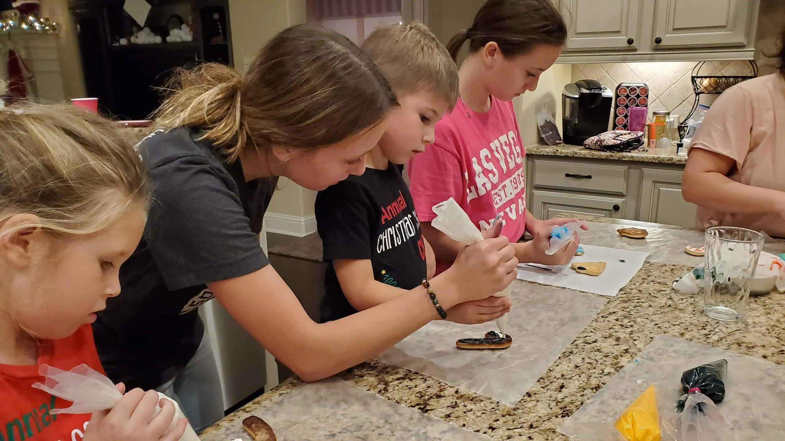 A group of children and a woman are gathered around a kitchen counter, decorating cookies with icing and festive toppings for the holidays.