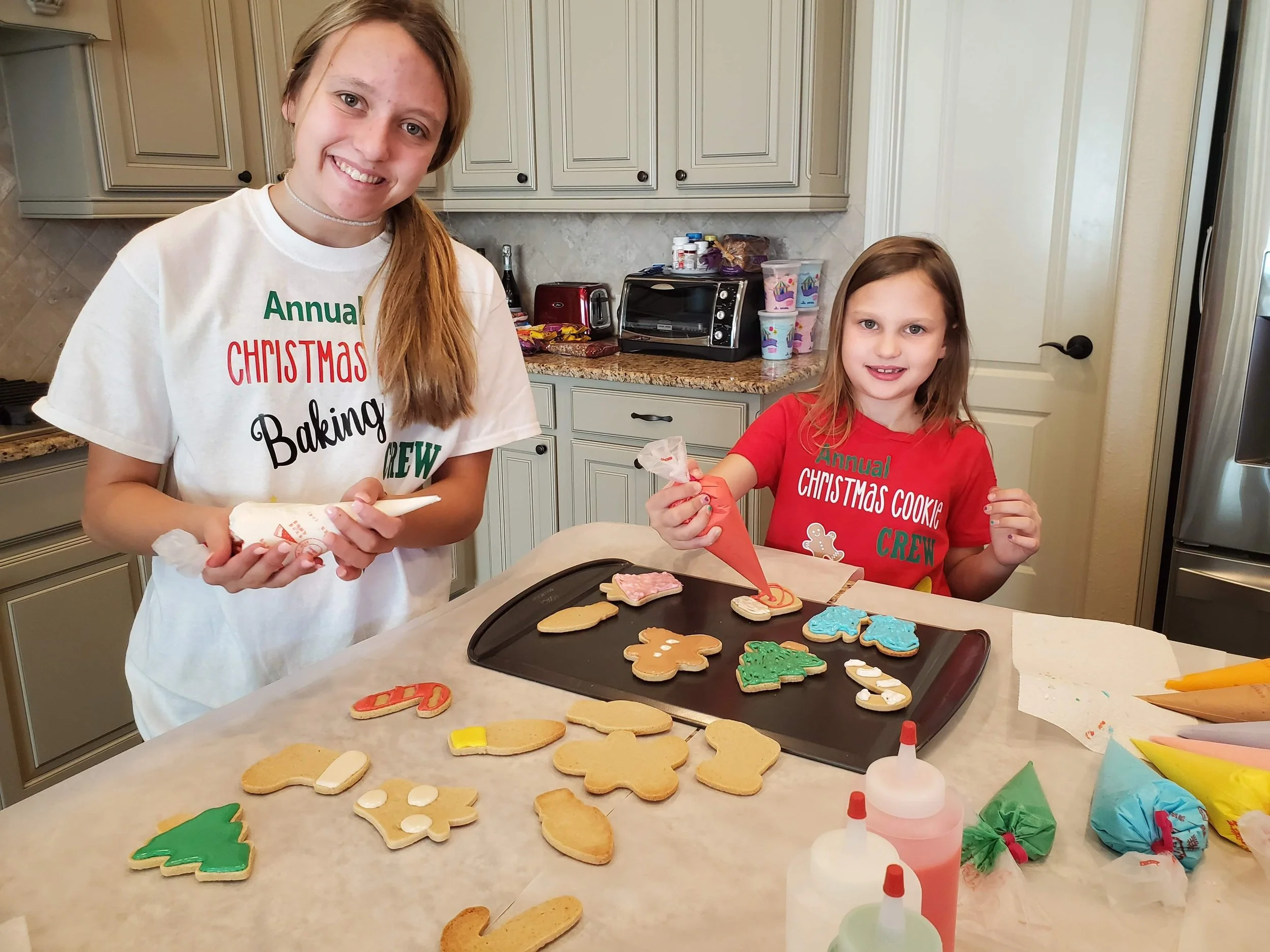 Two girls decorating Christmas cookies in a kitchen, with plates of decorated cookies on the counter.