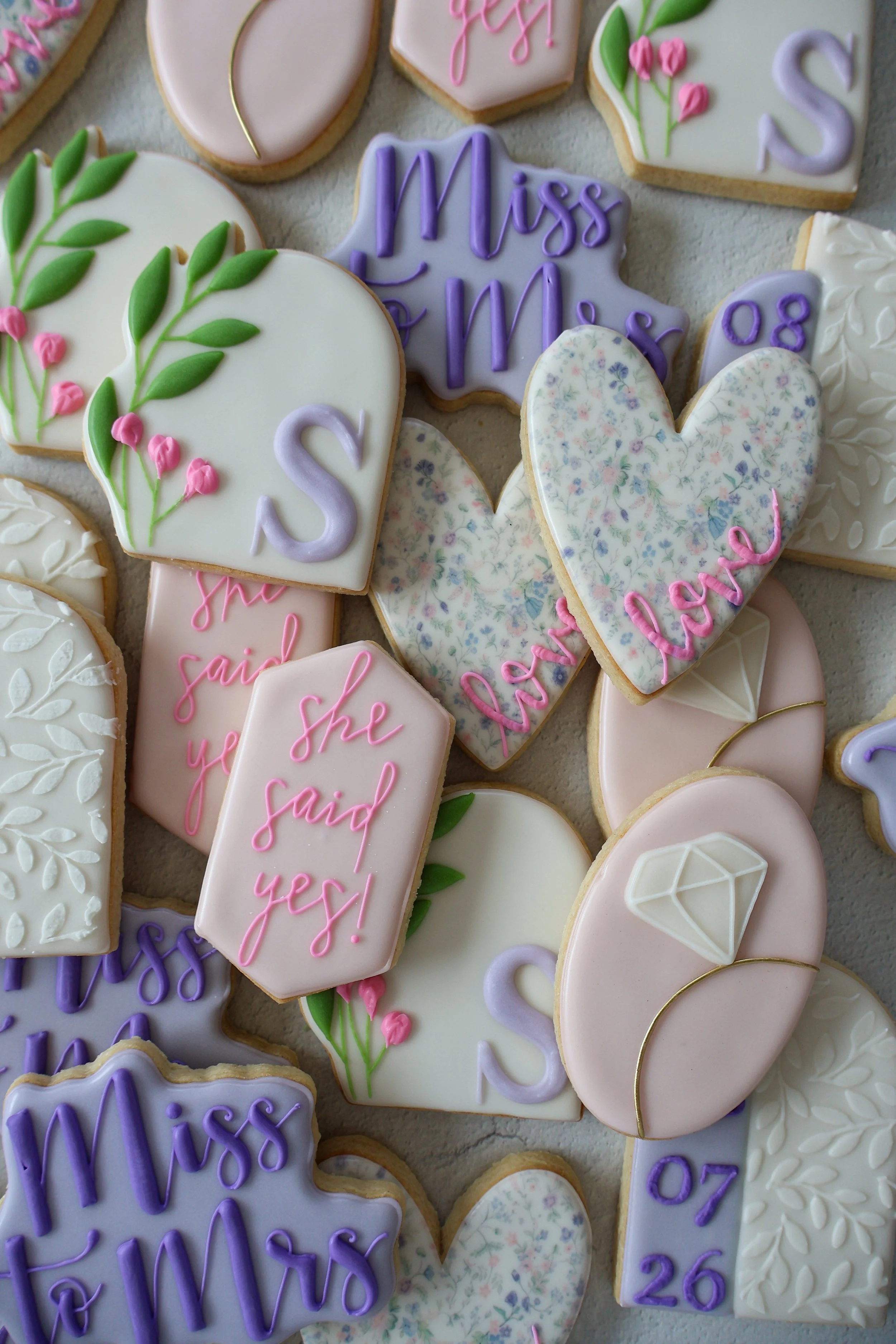 Assorted decorated cookies with floral and heart designs, some with phrases like "Miss" and "she said yes!" in pastel shades of pink, purple, and white.
