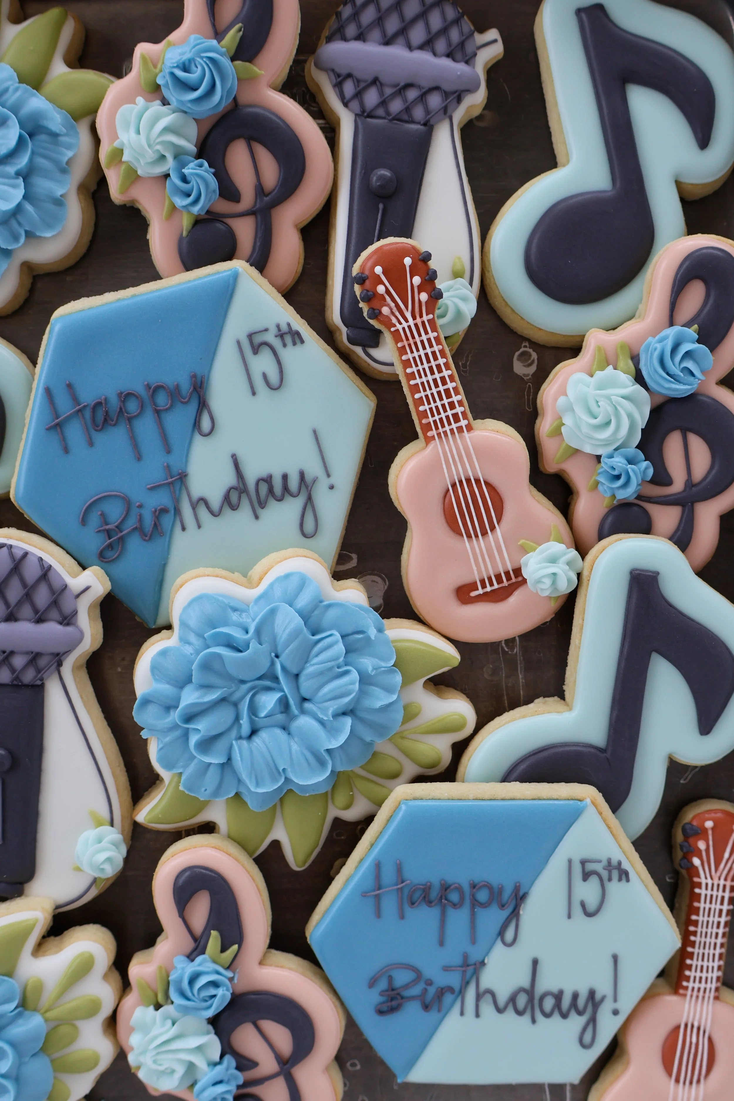 Collection of decorated cookies for a 15th birthday celebration, including a blue and light blue hexagon-shaped cookie with the message "Happy 15th Birthday!", a guitar cookie with pink, white, and brown icing, and various music-themed cookies shaped
