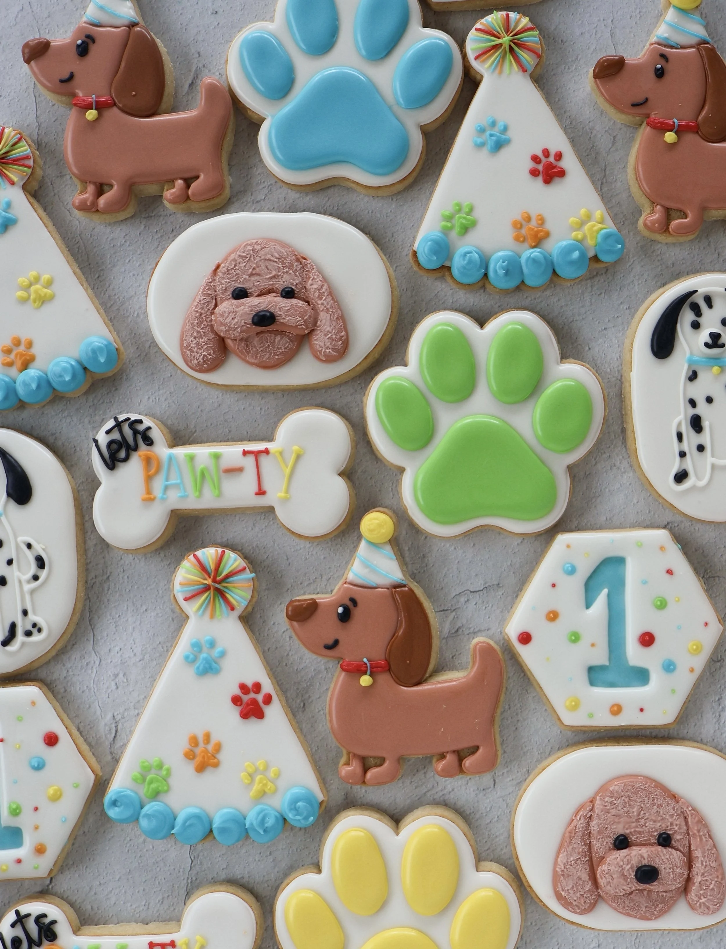 Assorted decorated cookies with dog and paw print themes, including puppy-shaped cookies, paw print designs, and birthday-related cookies with colorful icing and sprinkles.