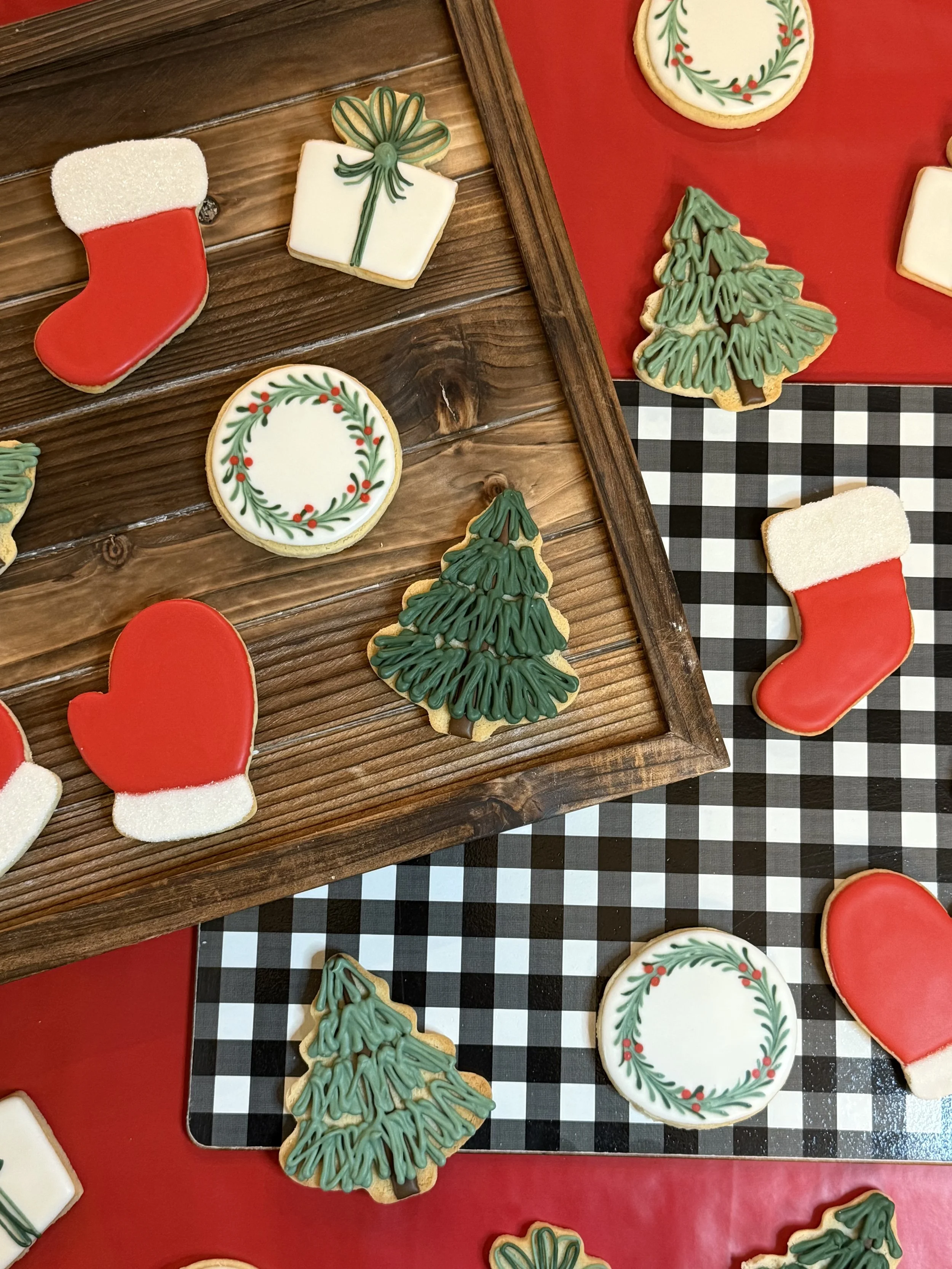 Assorted decorated Christmas cookies shaped like stockings, gift boxes, wreaths, and Christmas trees on a wooden and checkered surface.