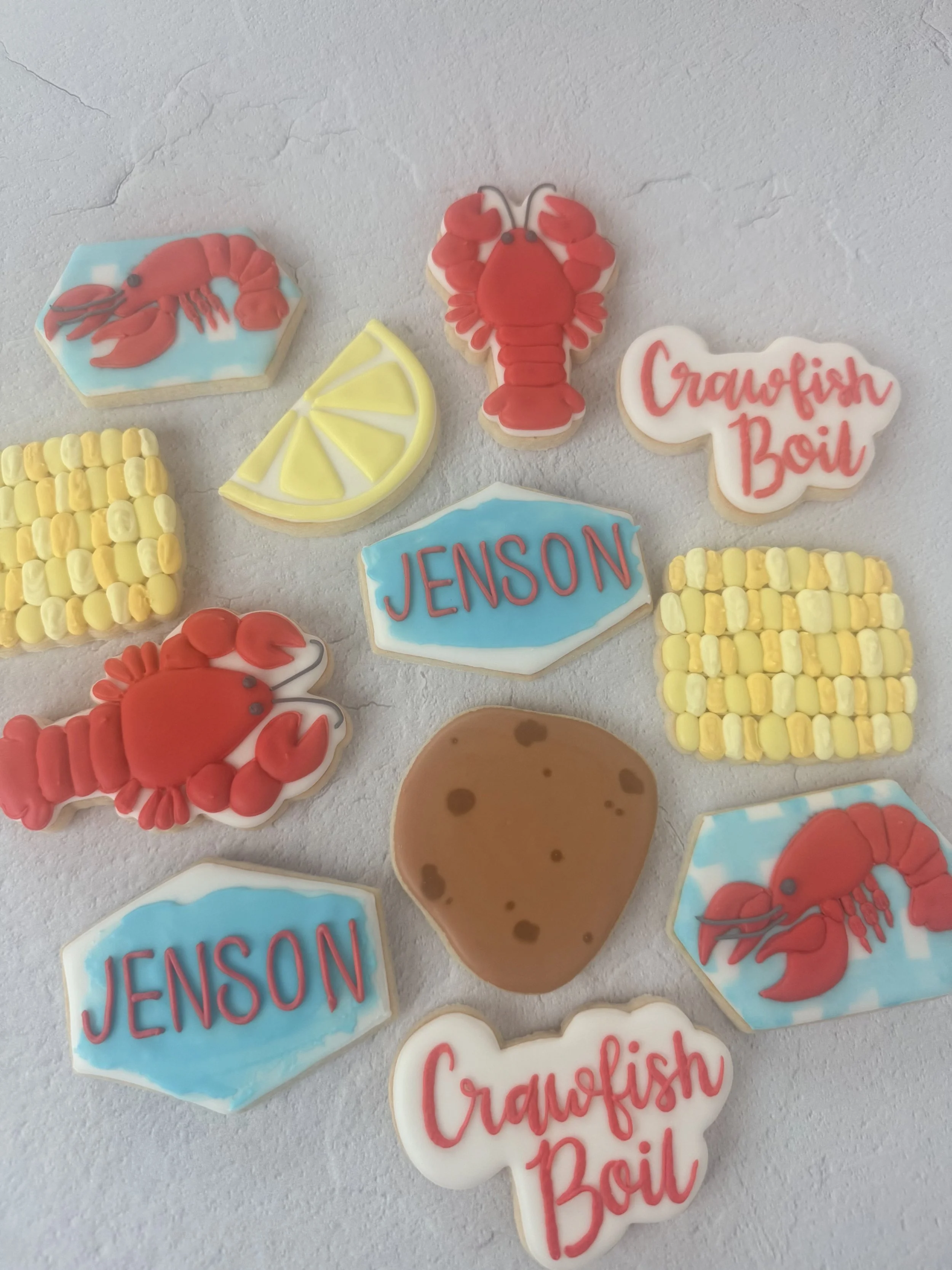 Collection of cookies shaped and decorated as a lobster, lemon wedge, a sign with the name 'Jenson', a sign saying 'Crayfish Boil', a cookie with yellow and white beaded pattern, and a cookie with the words 'Crayfish Boil' written in red icing.