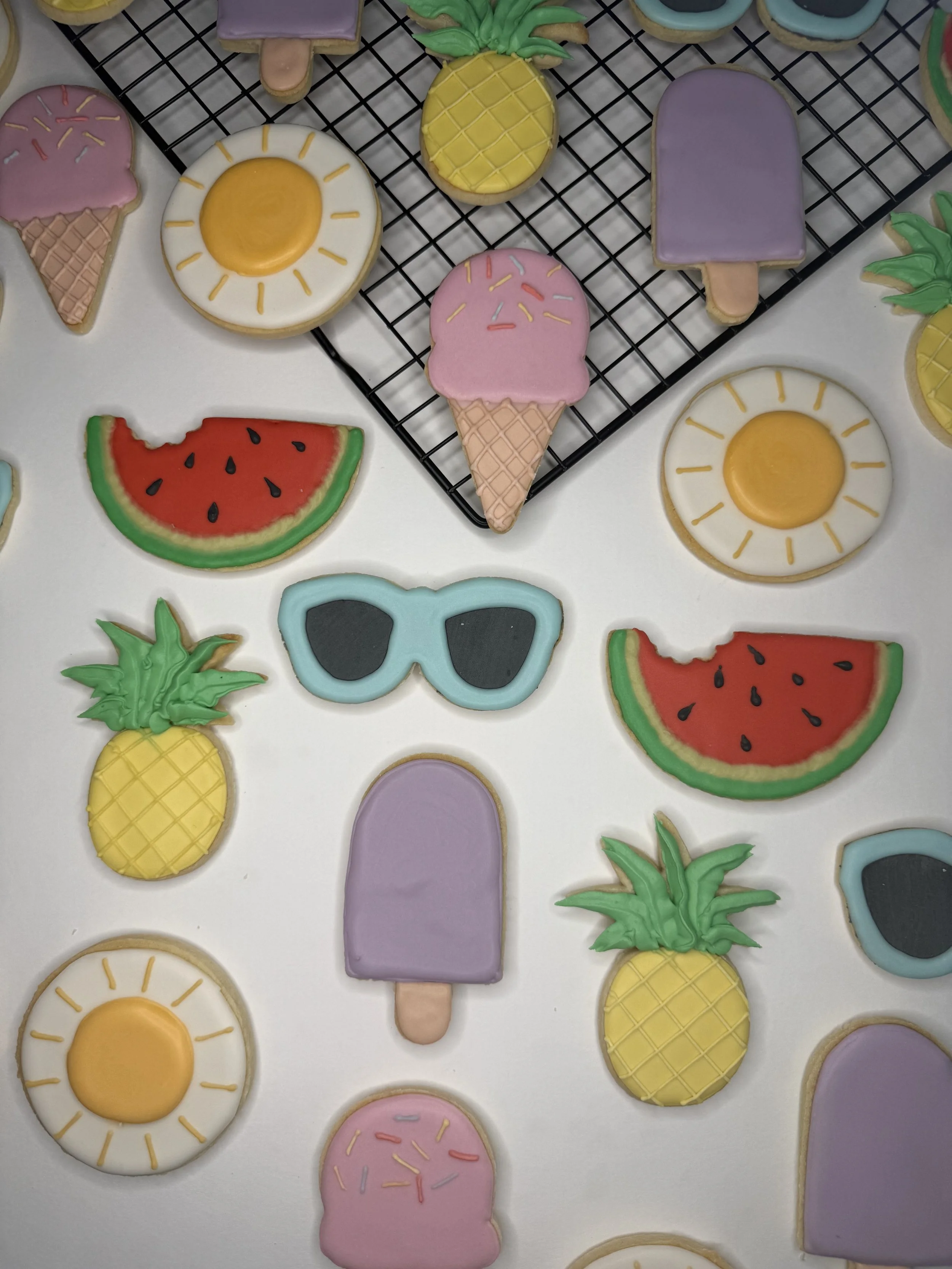Assorted colorful sugar cookies shaped like pineapples, watermelons, sunglasses, popsicles, pizza slices, and eggs, decorated with pastel and bright icing, placed on a white surface and a black cooling rack.