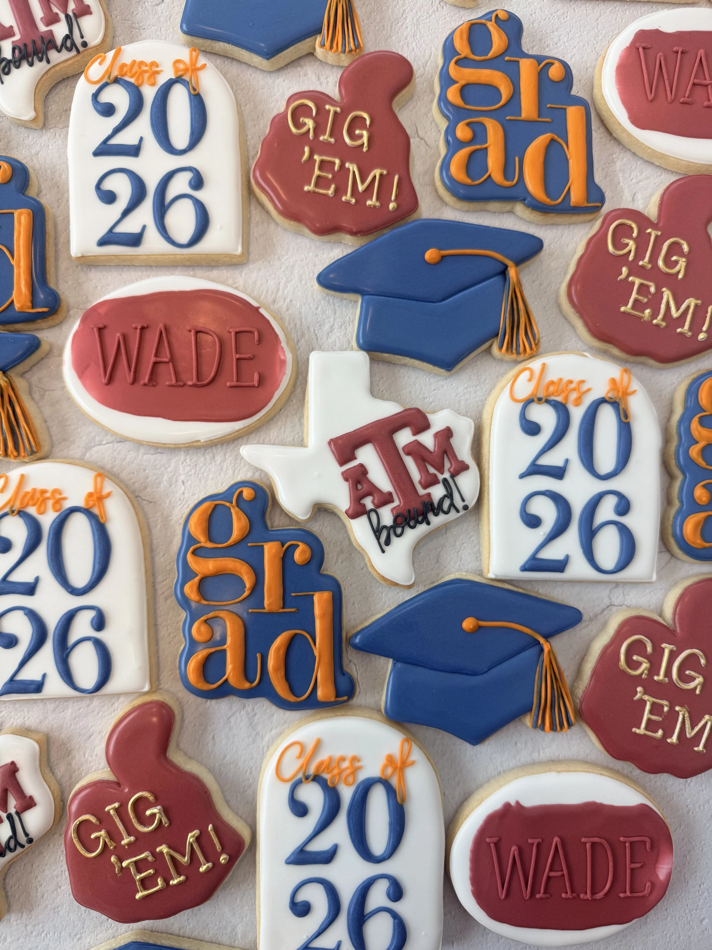 Collection of decorated sugar cookies with graduation themes. The cookies are shaped like graduation caps, diplomas, and speech bubbles. Colors include blue, red, white, and orange, with phrases like "Class of 2026," "GIG EM!" and "Wade."