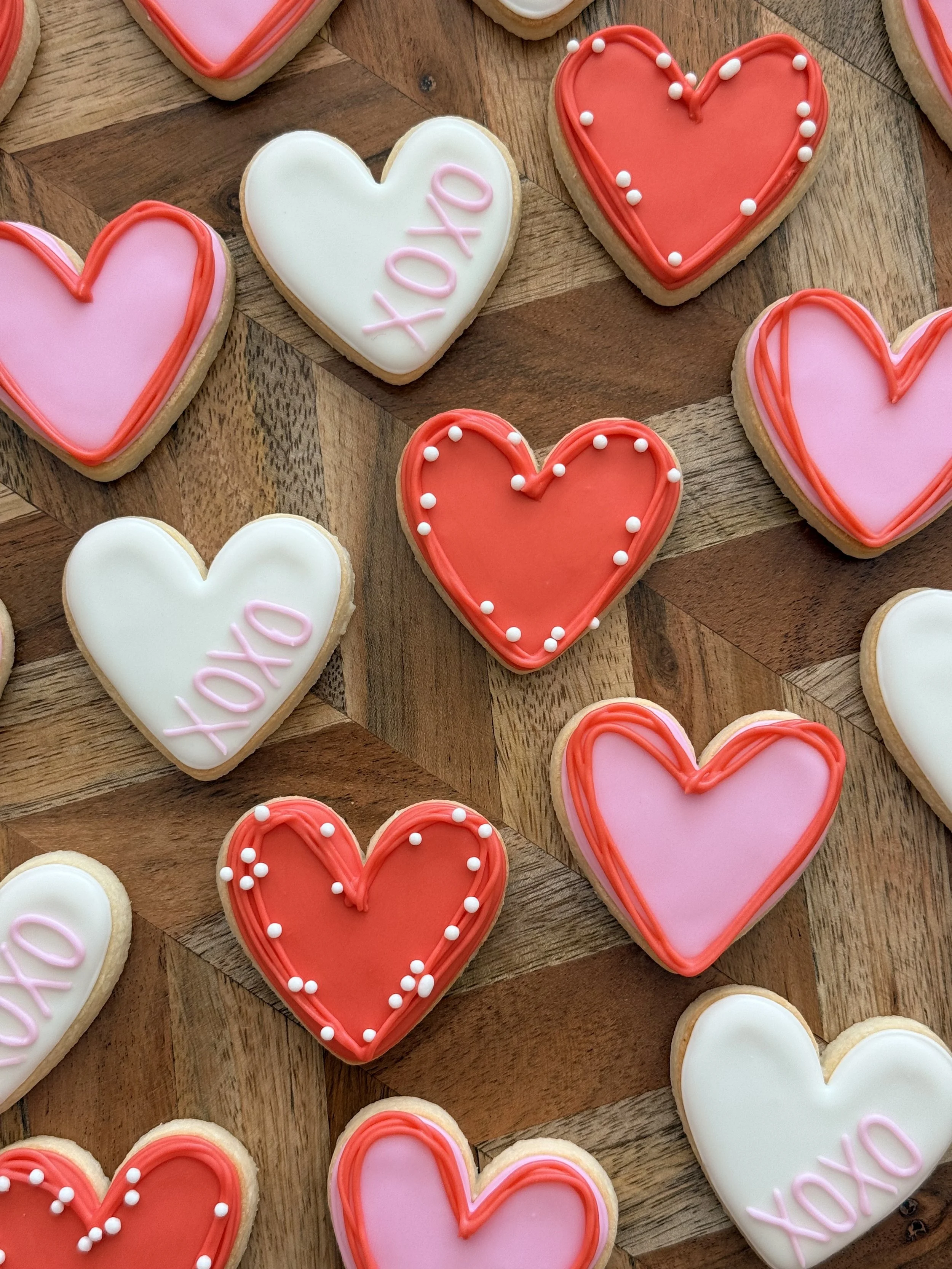 Heart-shaped cookies decorated with pink, white, and red icing, some with the word 'XOXO' and others with dotted and lined borders, arranged on a wooden surface.