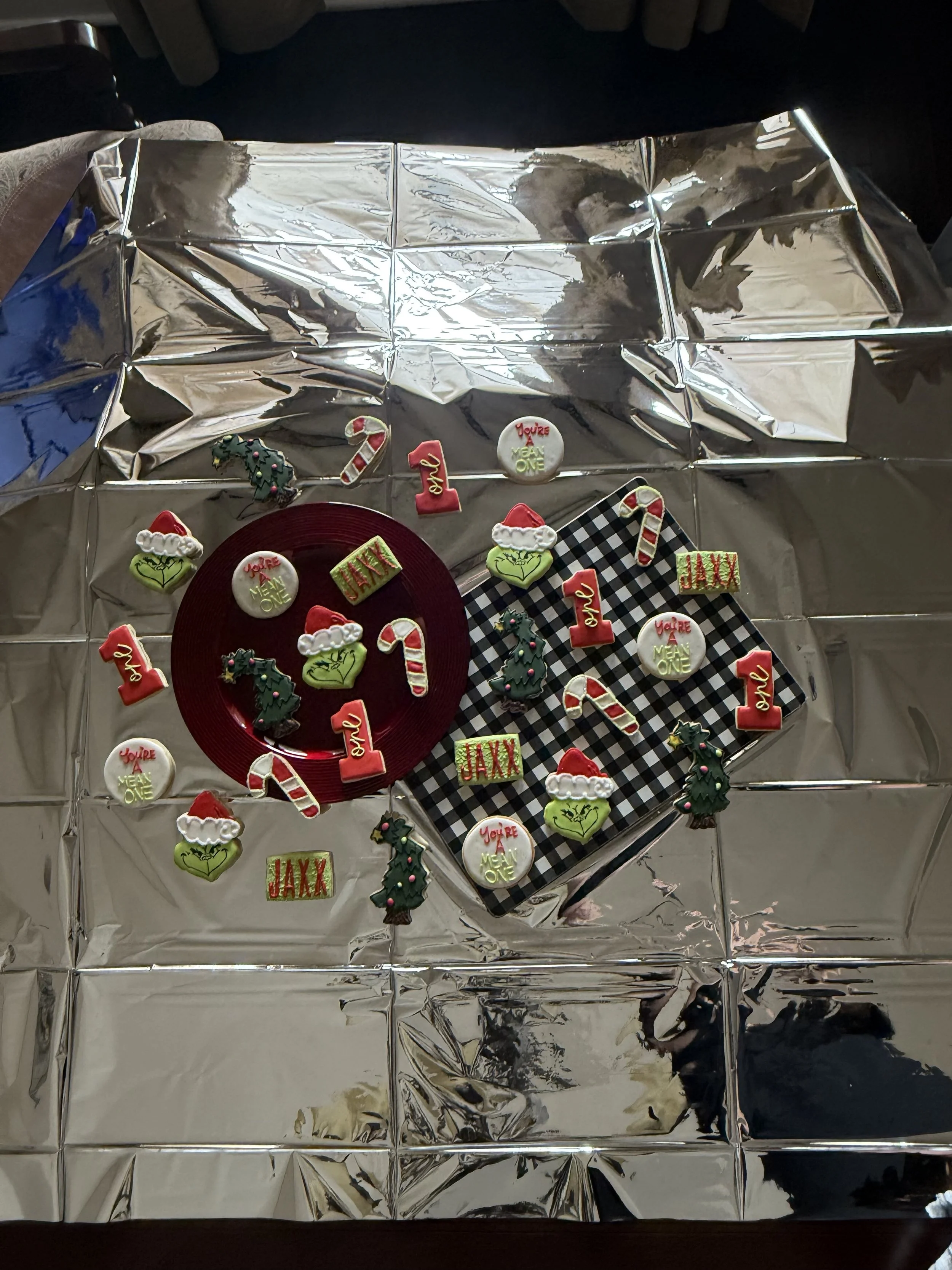 Holiday table setup with a silver foil tablecloth, decorated cookies shaped as holiday symbols and words such as 'JAXX' and 'you're a mean one', a red plate with cookies on top, and black and white checkered and plain plates.