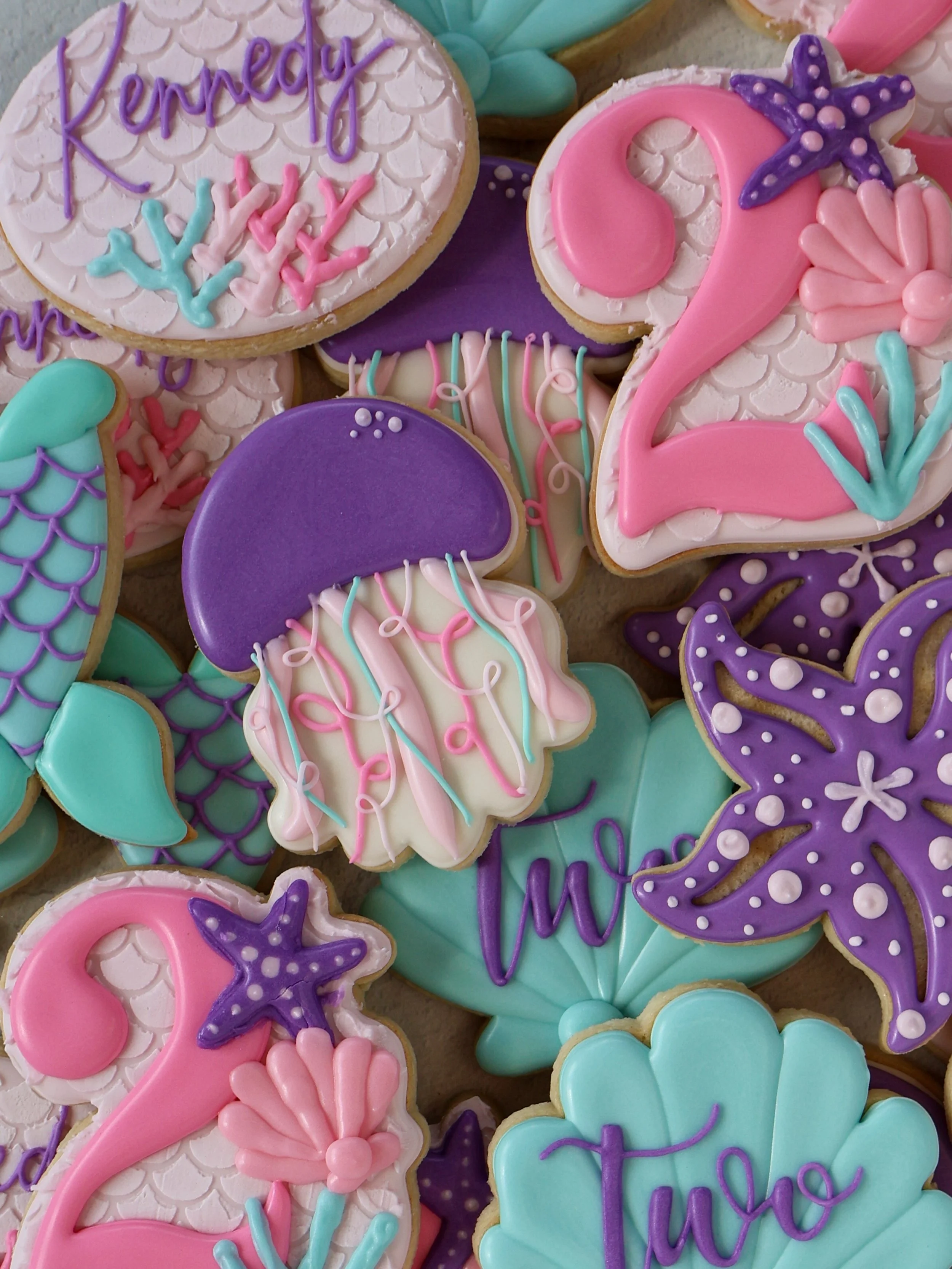 Colorful decorated cookies for a celebration, including a cookie with the name "Kelsey" written on it, and various other designs such as seashells, stars, hearts, and the number "2" in pink, purple, blue, and white icing.