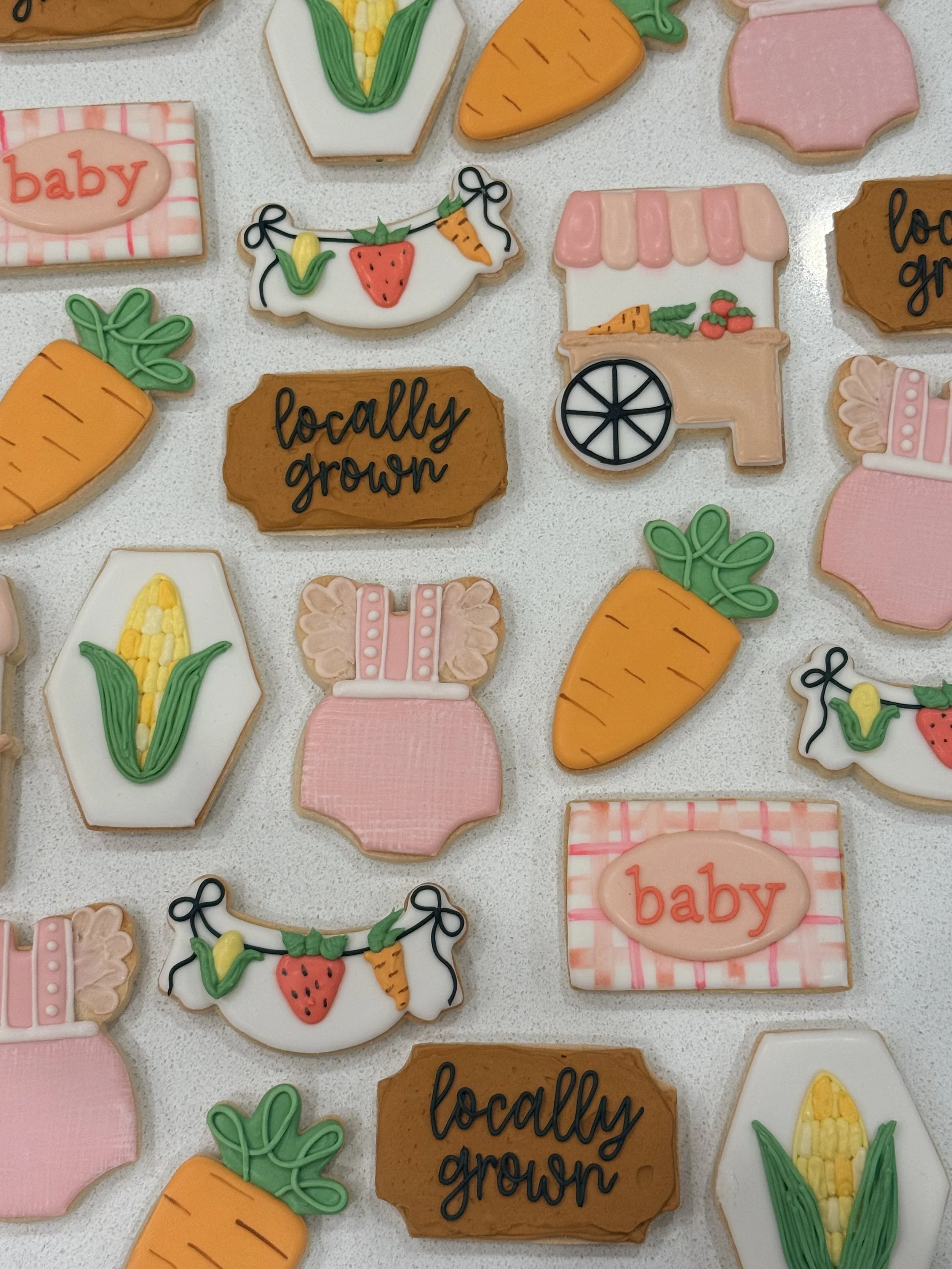 Assorted decorated cookies shaped like carrots, strawberries, pink cakes, a shopping cart, corn on the cob, and signs with words like 'baby' and 'locally grown.'