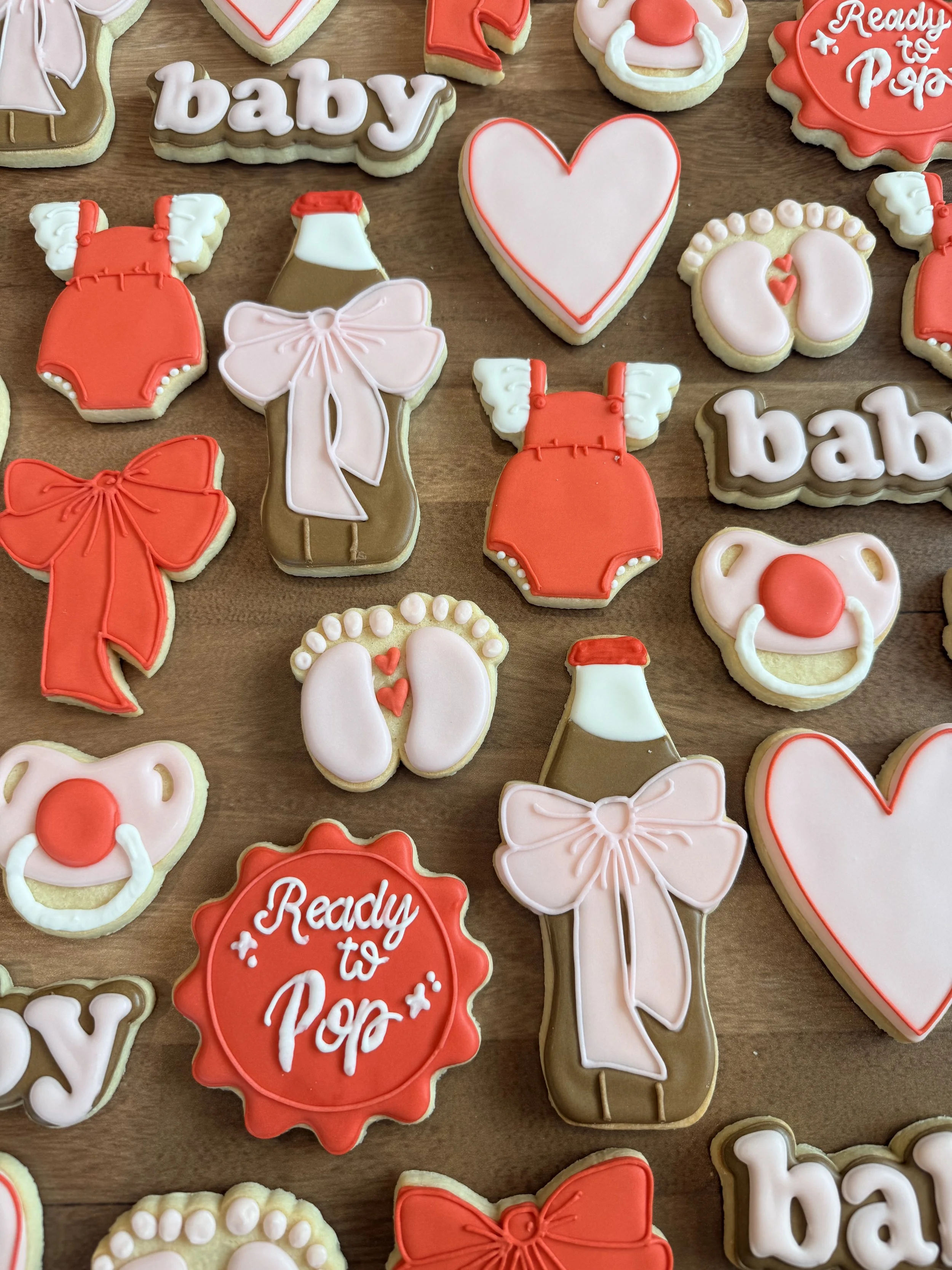 A collection of decorated cookies on a wooden surface, including cookies with the words 'baby' and 'ready to pop,' cookies shaped like hearts, baby bottles, pacifiers, and bows, all decorated with pink, white, brown, and red icing.