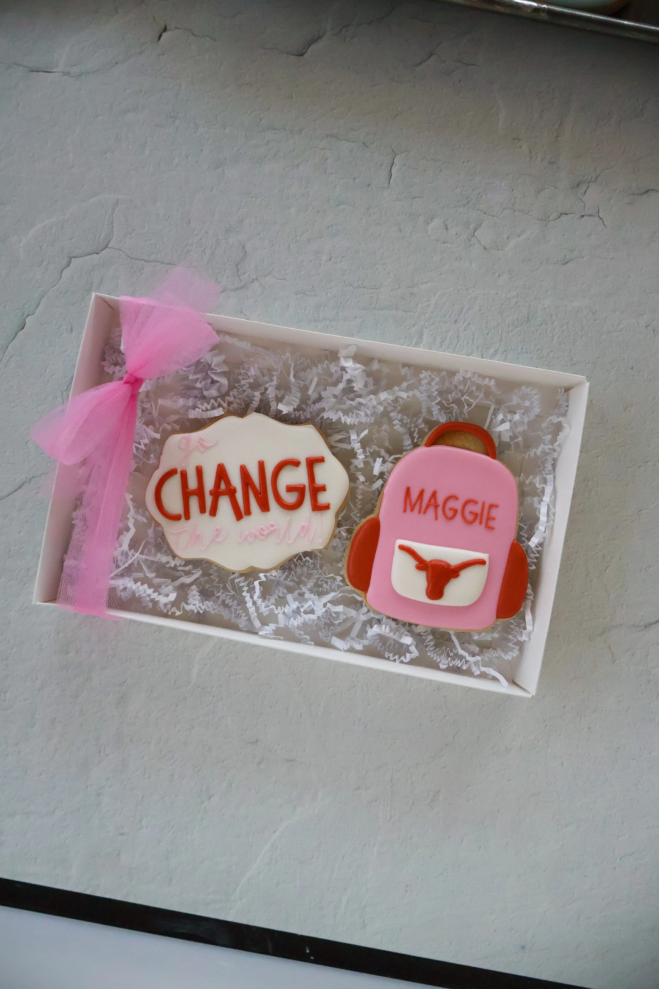 Decorated sugar cookies in a white box with crinkled white paper filling. One cookie has a white icing with the words 'CHANGE' and pink accents, and another cookie is shaped like a pink backpack with the name 'Maggie' and a longhorn symbol.
