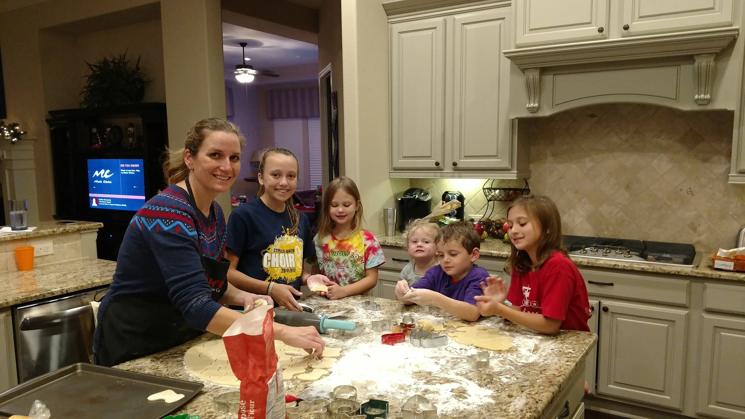 A woman and five children in a kitchen baking cookies together. The woman is smiling and wearing a black apron. The children are gathered around a countertop with baking supplies and cookie cutters, engaged in baking activities.