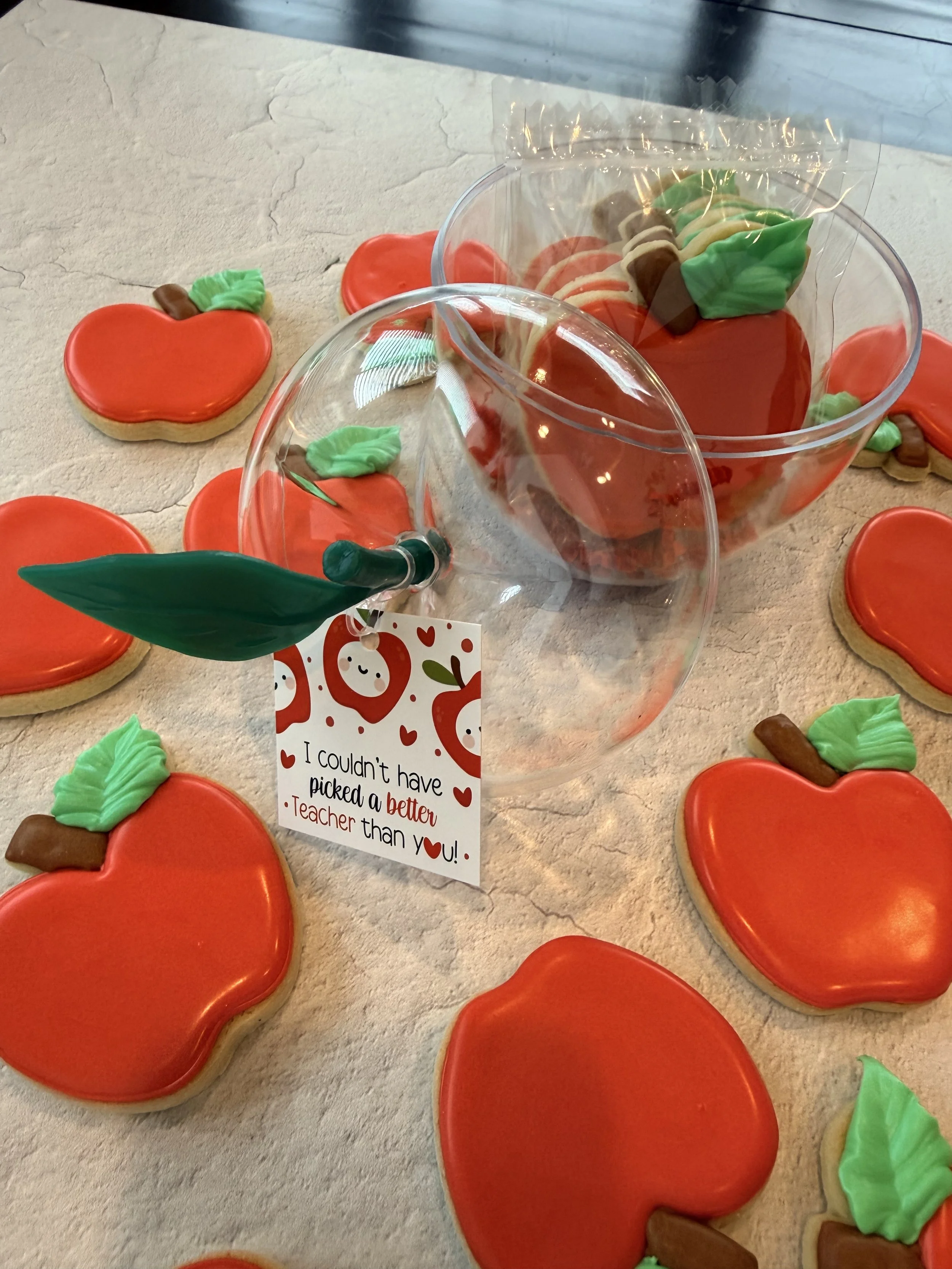 Colorful apple-shaped cookies with green leaves, red icing, and brown stems are arranged on a beige textured surface. Large glass bowls containing more of these cookies are also present. There is a partially visible card with a holiday or love theme 