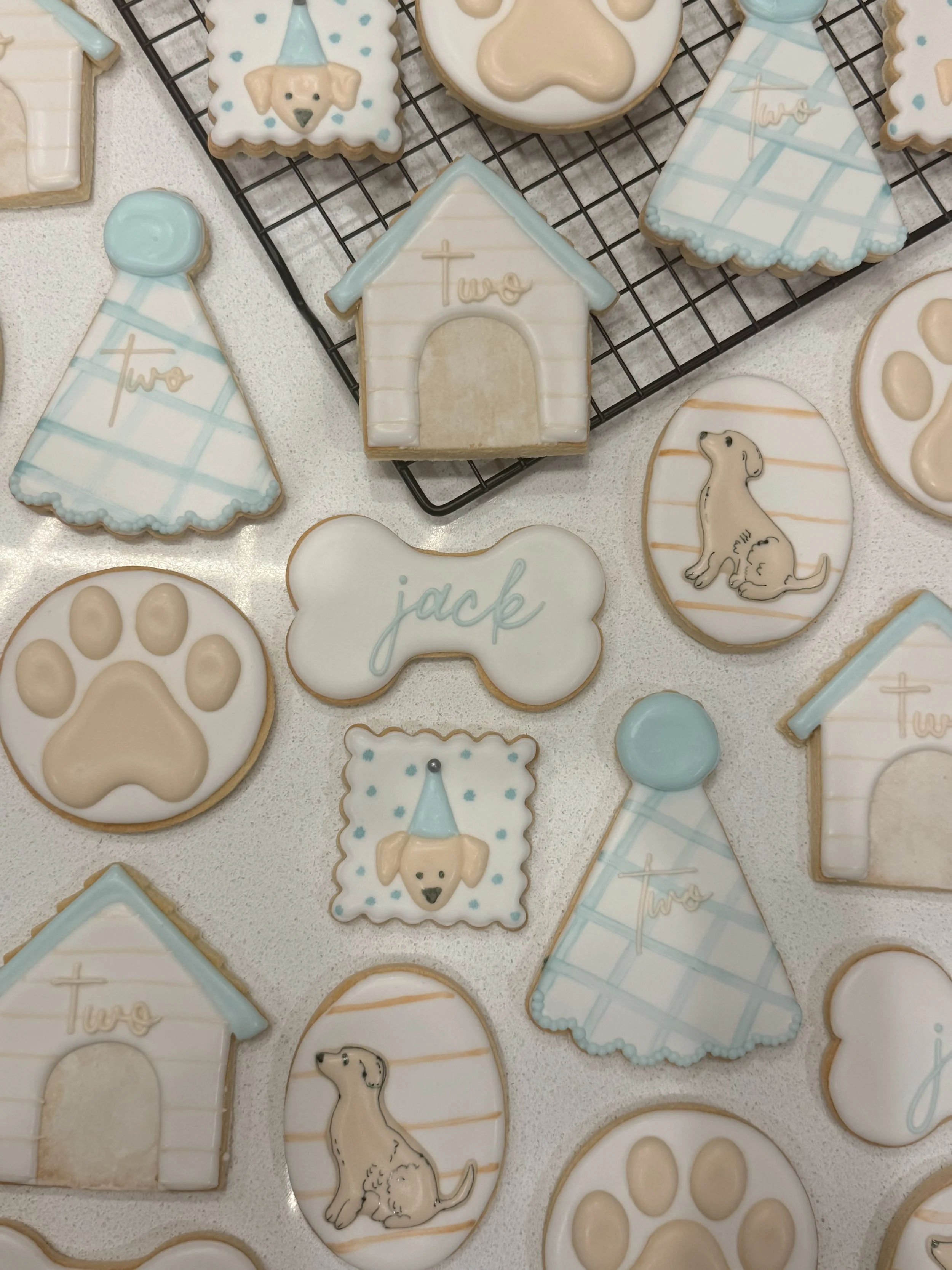 Assorted decorated cookies with themes of dogs, bones, dog houses, and the words 'two' and 'jack', in pastel blue, white, and beige colors.
