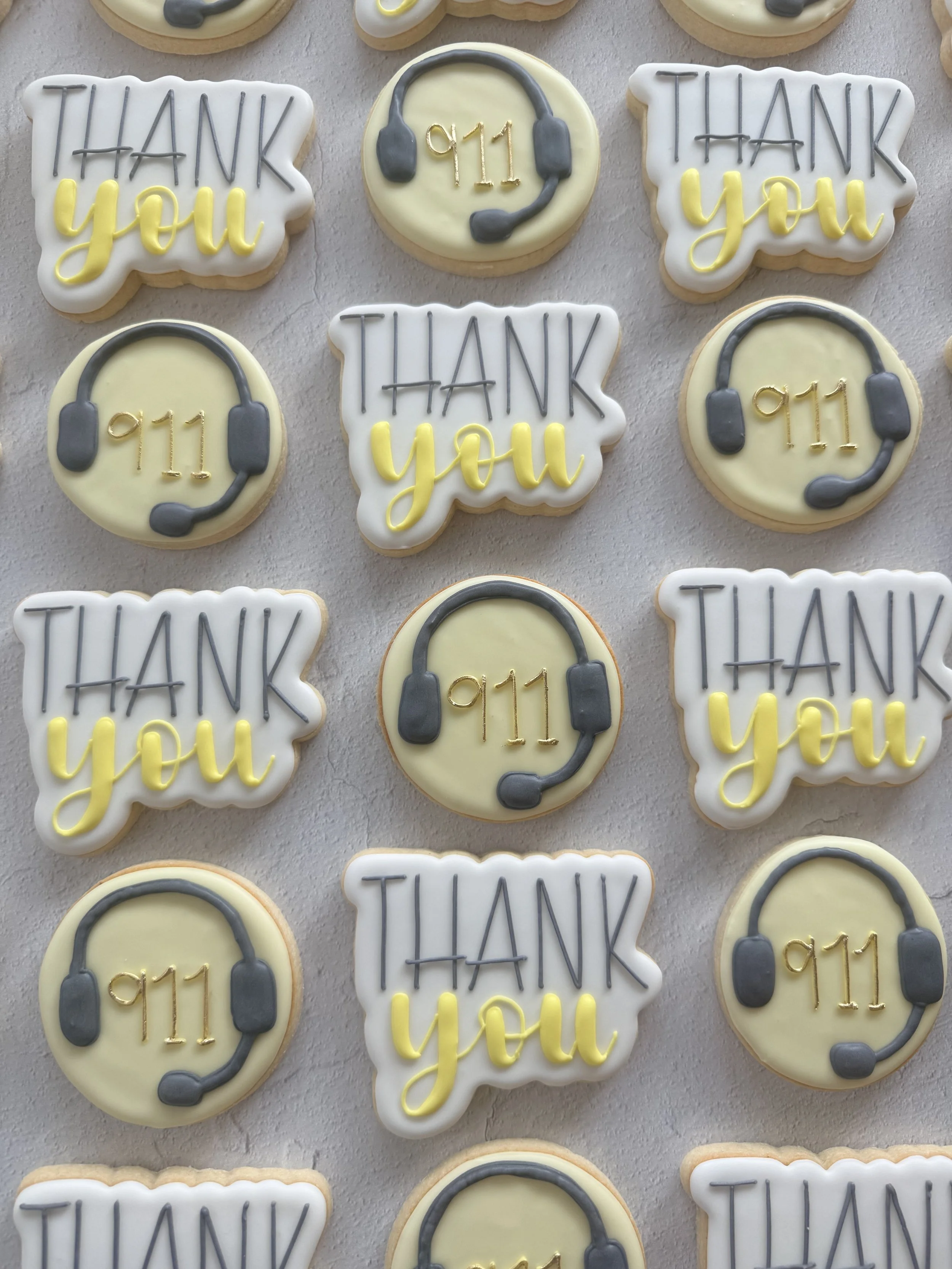Cookies decorated with messages of appreciation: some cookies feature the words "Thank You" in black and yellow icing, and others depict a headset with the numbers "911" in gold icing, all on a light surface.