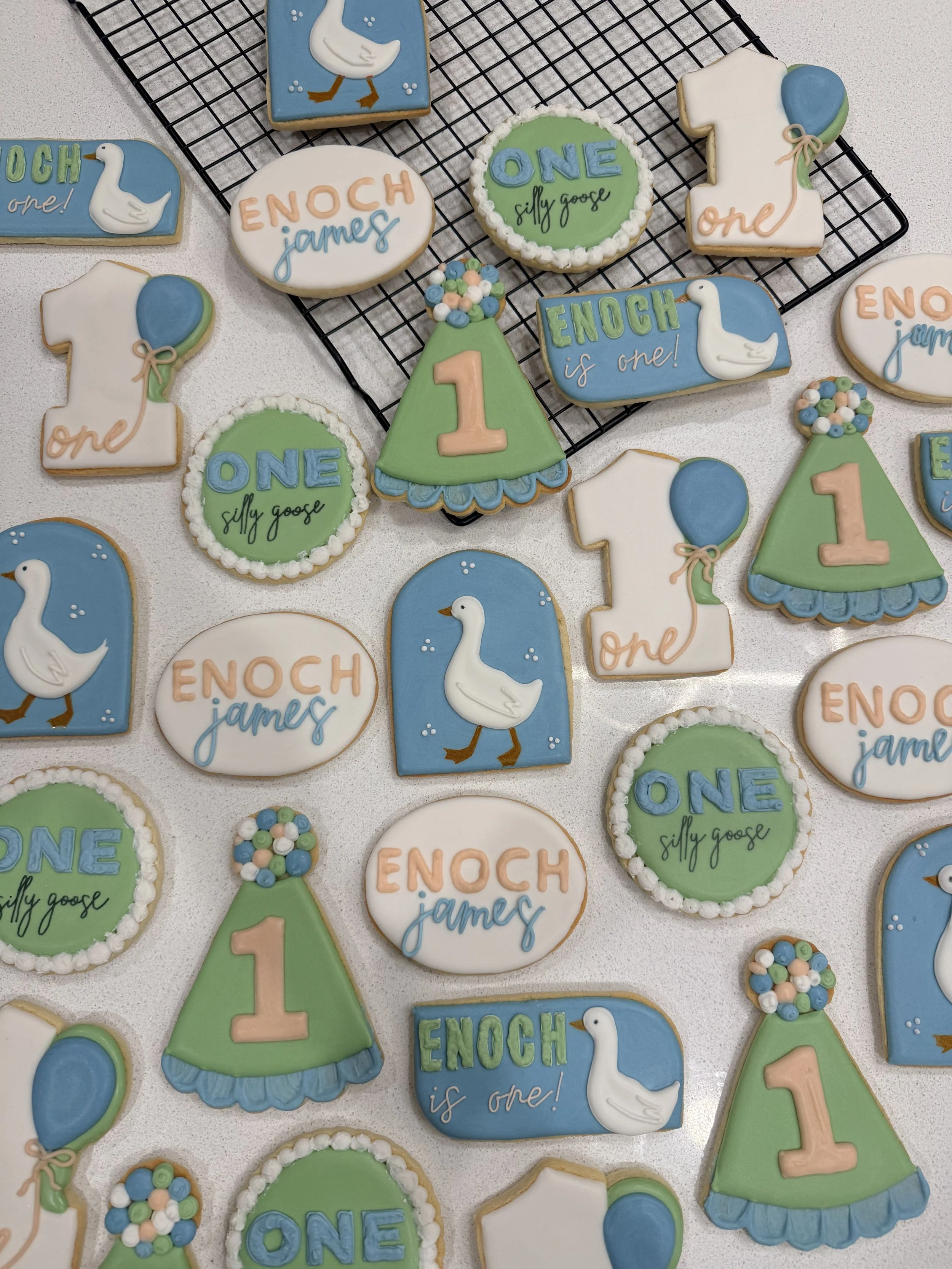 Assorted cookies with baby shower themes, including ones with swan, balloon, and number '1' designs, decorated with pastel colors like blue, green, white, and peach.