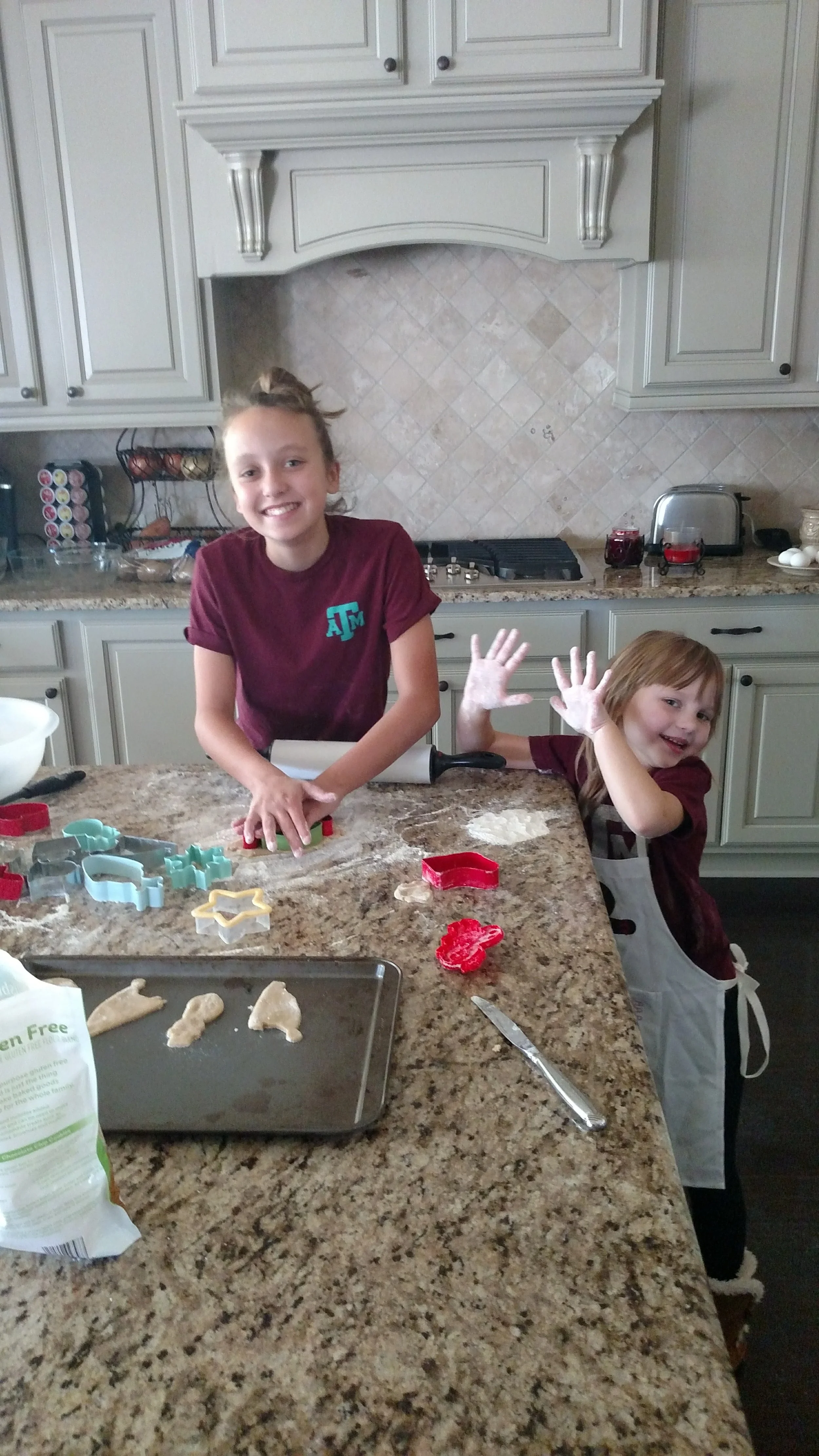 Two young girls in a kitchen, making cookies with cookie cutters on a granite countertop. One girl is smiling at the camera, the other is waving and smiling.