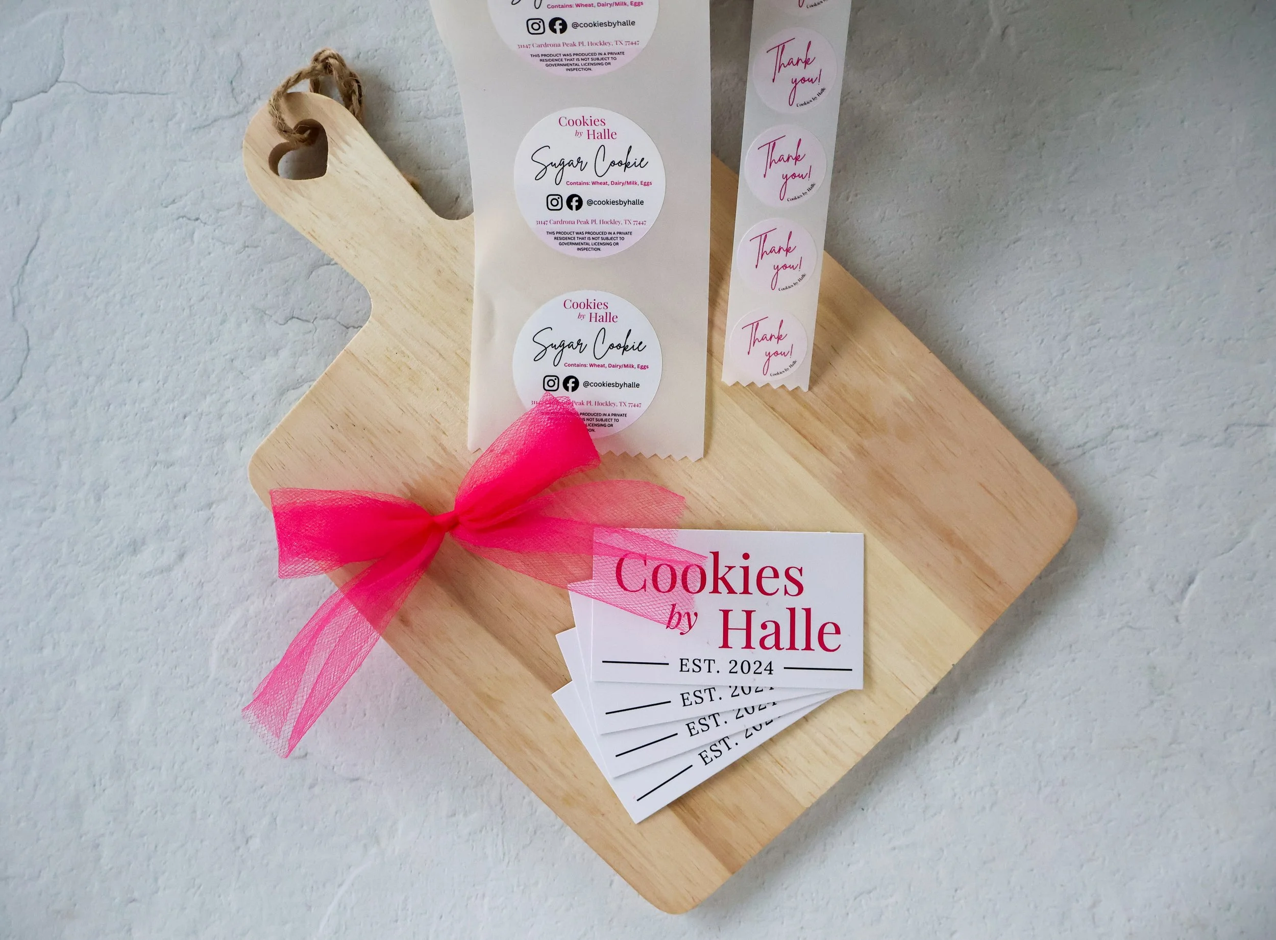 A wooden cutting board with a pile of business cards from Cookies by Halle, featuring pink, black, and white design elements, along with a roll of thank you stickers with pink writing, and a pink ribbon tied in a bow.