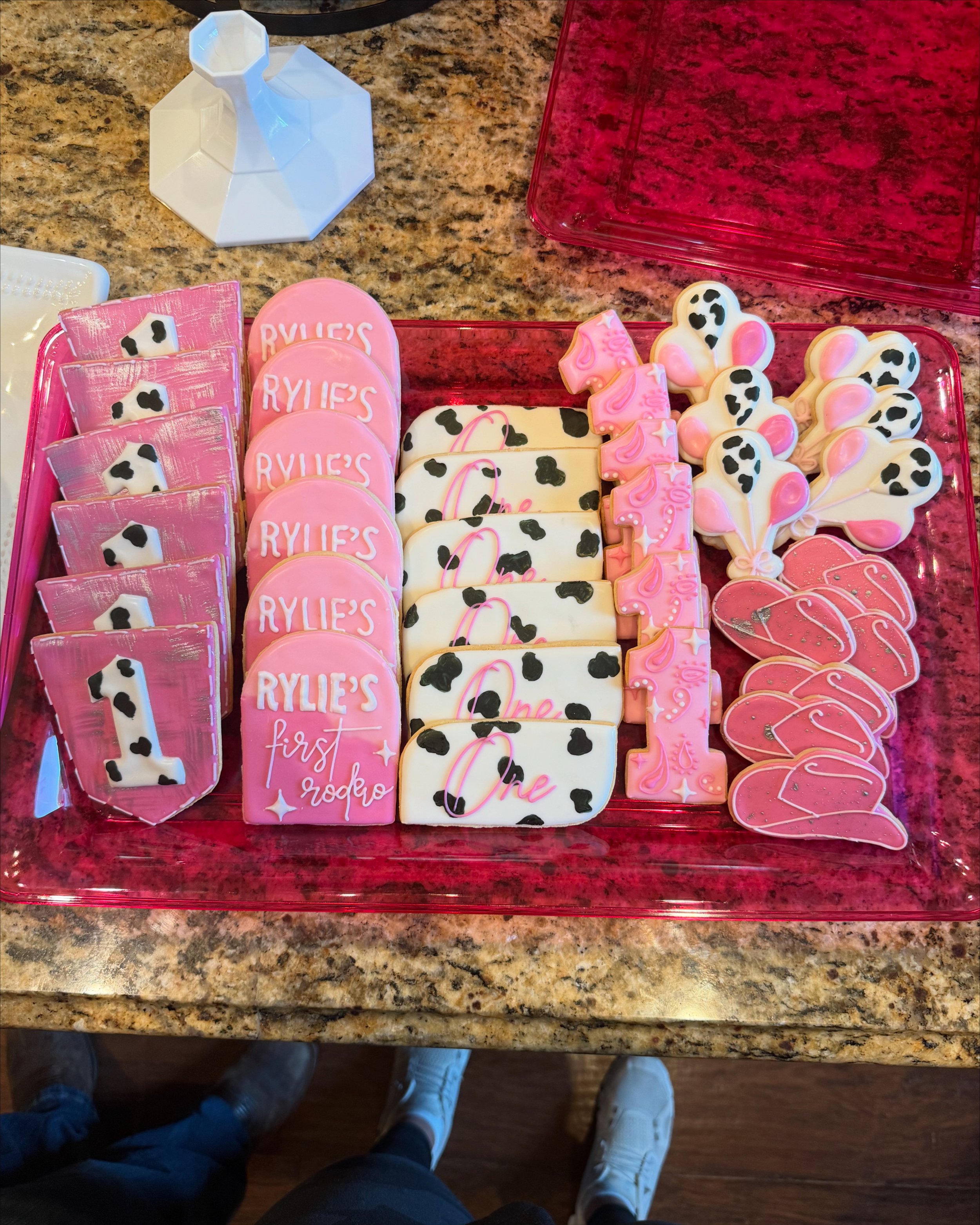 A tray of decorated cookies with pink, white, and black designs, including cow spots, hearts, the number one, and writing. Visible on the cookies are phrases like 'RYLIE'S first' and 'One'. The tray is on a multicolored granite countertop, and part o