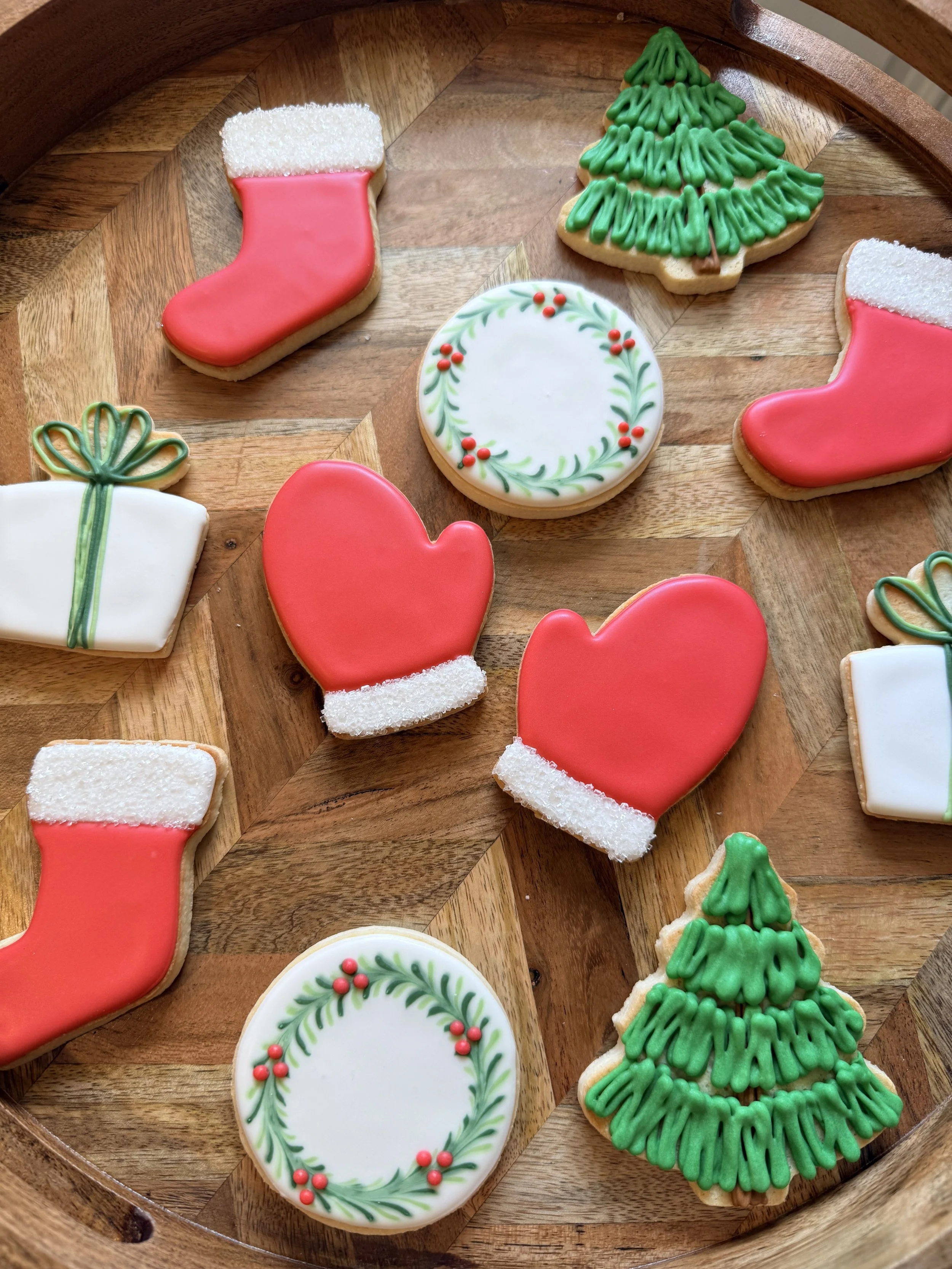 Decorative Christmas cookies on a wooden tray, including a Christmas stocking, Christmas tree, a round cookie with holly and berry design, and a gift box.