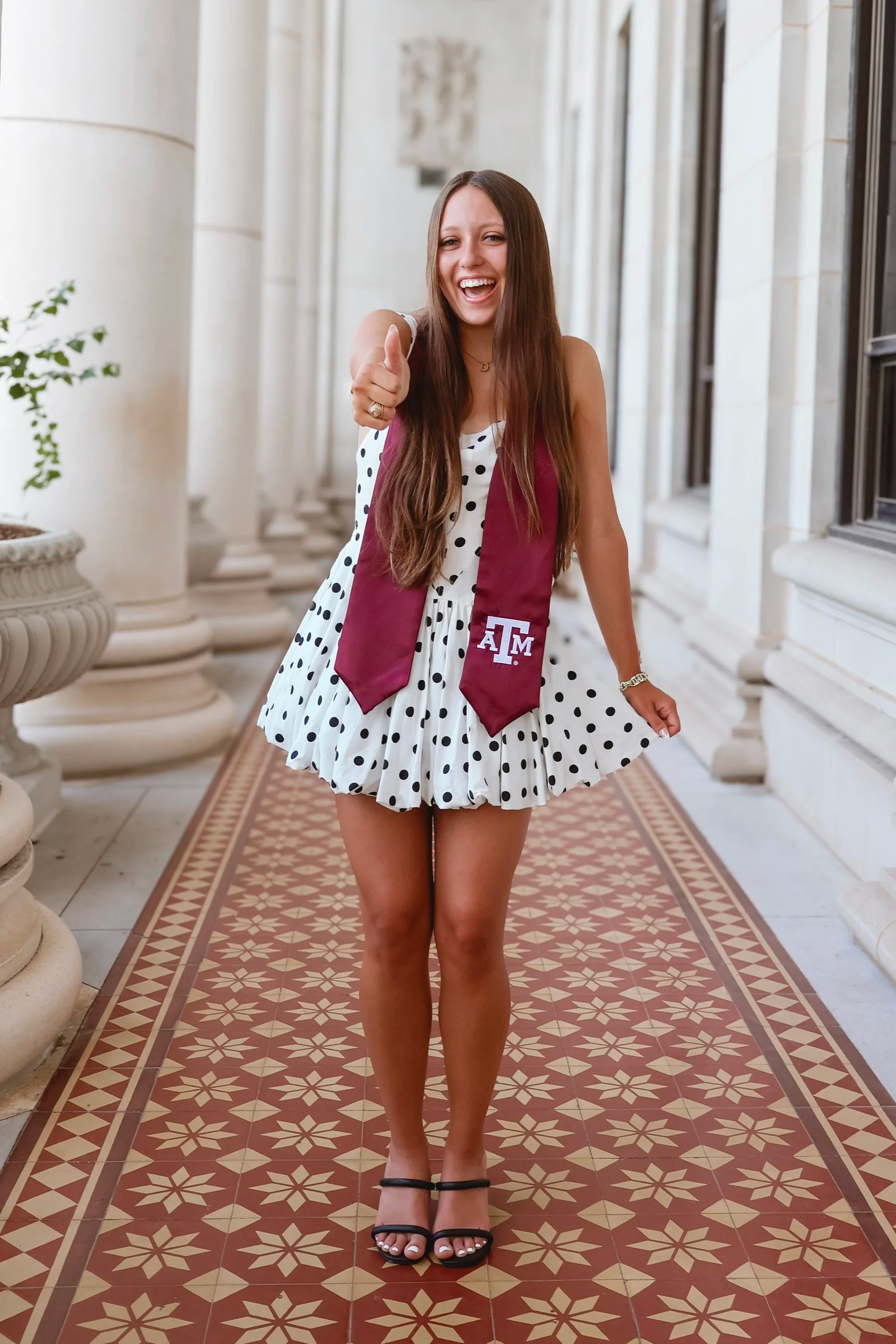 Young woman at graduation wearing a maroon and white dress with a maroon stole featuring the Texas A&M logo, standing on a patterned tile walkway with columns and windows in the background, smiling and pointing at the camera.
