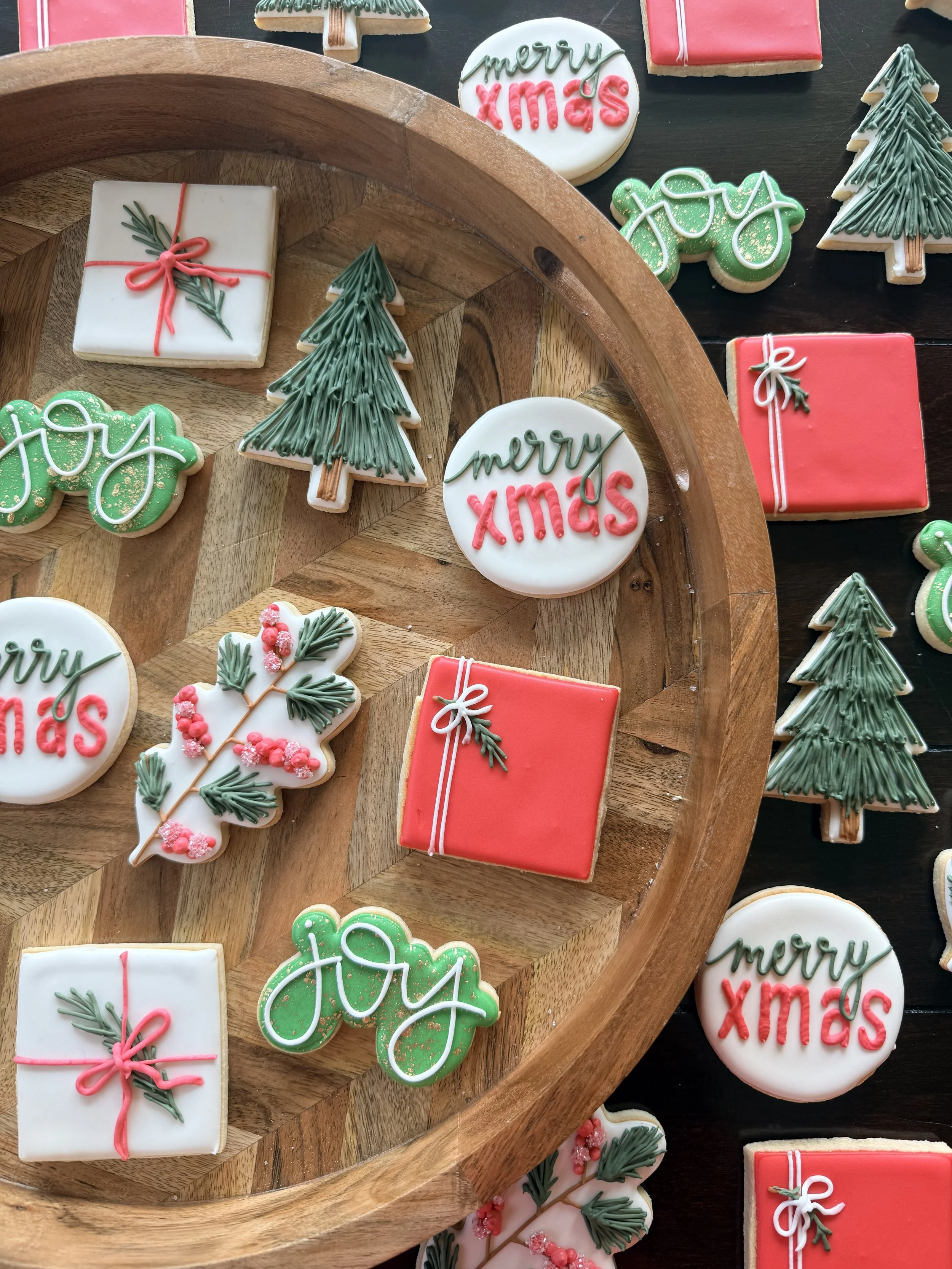 Various Christmas-themed decorated cookies, including gifts, trees, and a round cookie with 'Merry Xmas' written on it, all arranged on a wooden tray and dark surface.