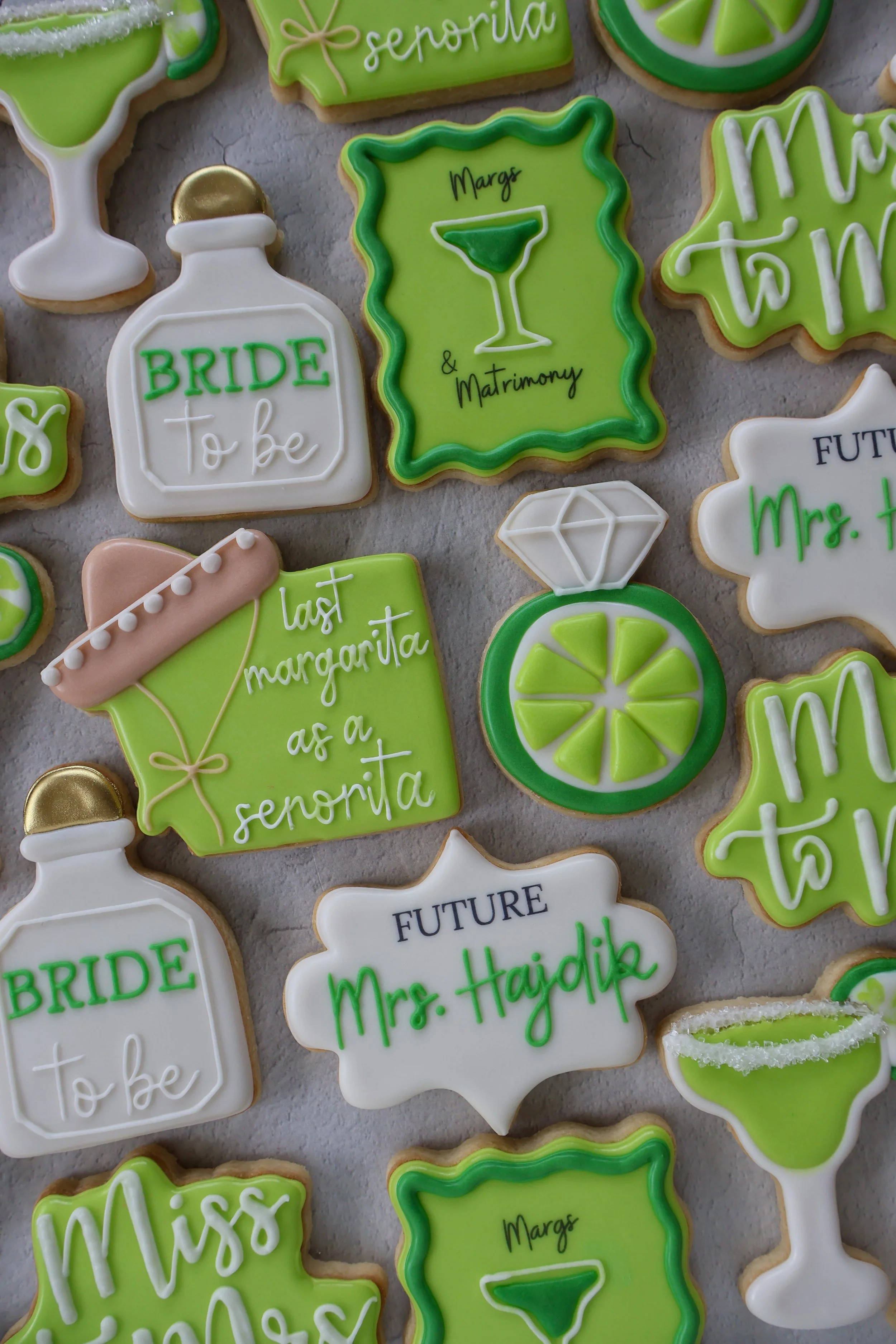 Decorated cookies with green, white, and gold icing, including phrases like "Bride to be," "Margs & Matrimony," and "Mrs. H" with lime slices and cocktail glasses.