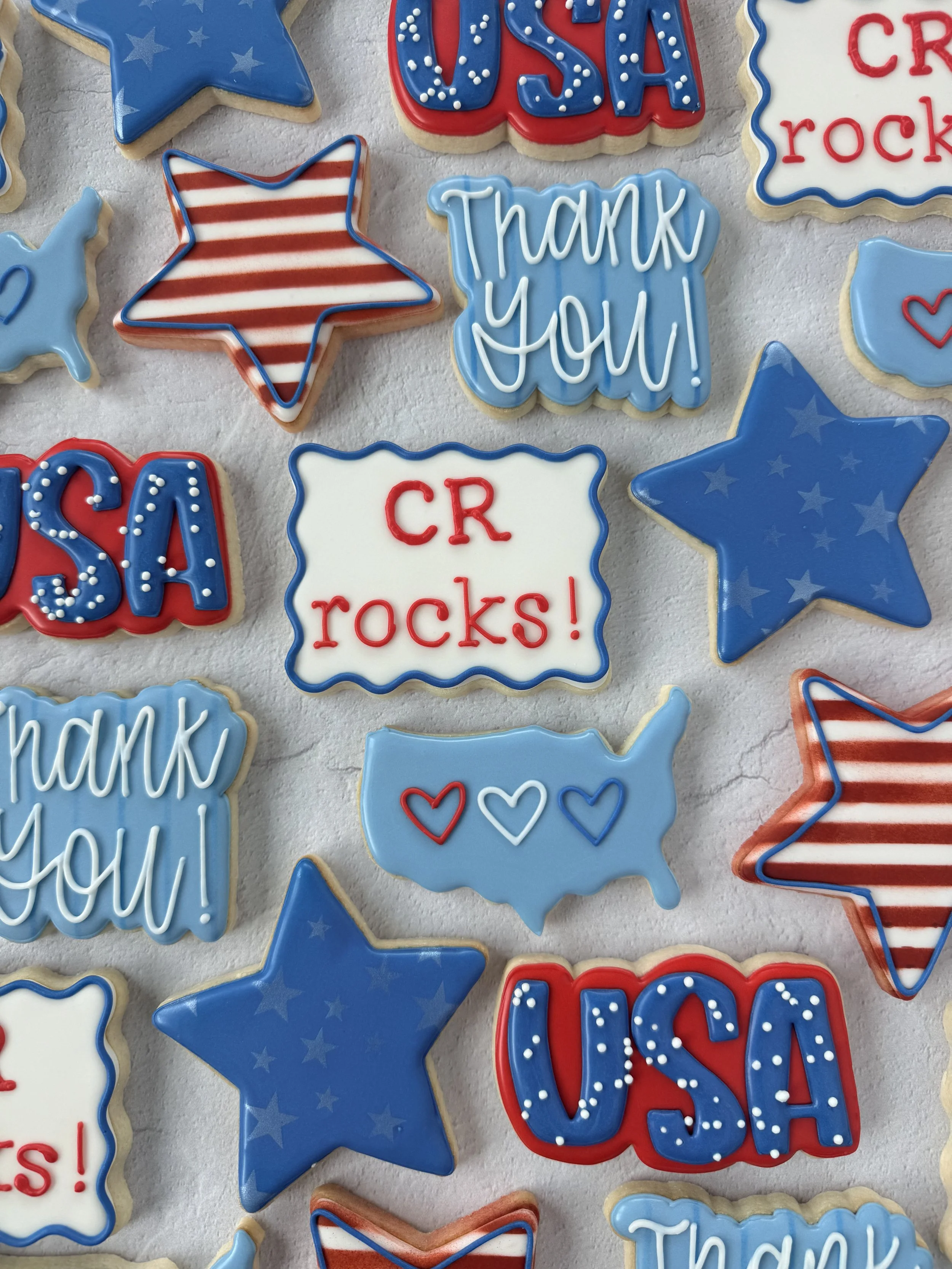 Decorative cookies with patriotic and thank you messages, shaped like stars, hearts, and the state of Texas, decorated with red, white, and blue icing.