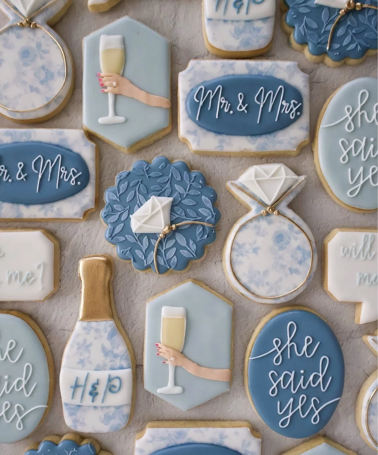 Decorative cookies with a wedding or celebration theme in blue, white, and gold colors. Some cookies have phrases like 'Mr. & Mrs.' and 'she said yes,' along with images of champagne bottles, hand holding a champagne glass, and diamond rings.