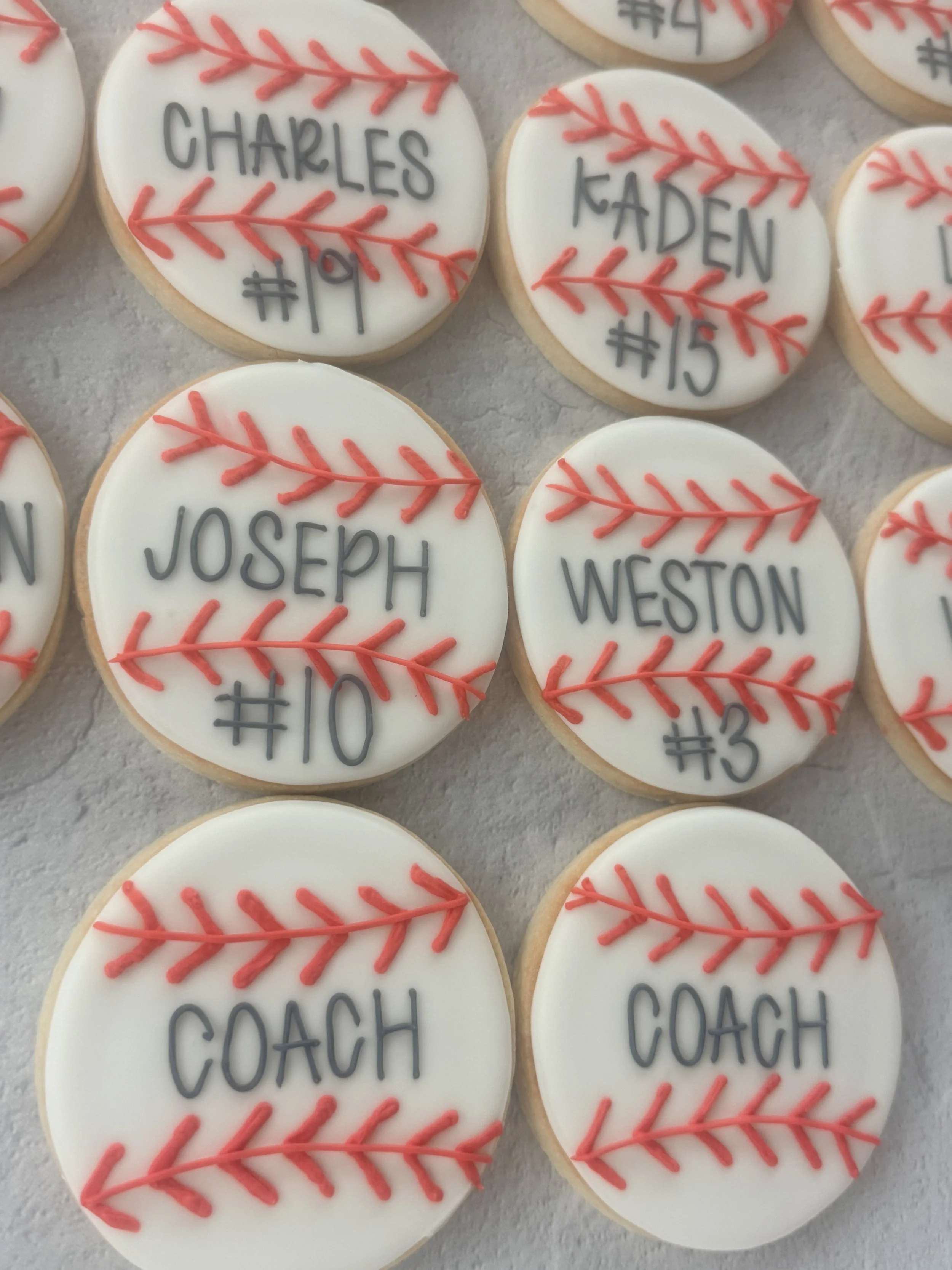 Decorated baseball-themed cookies with names and numbers written in black, featuring red icing for the baseball stitching.