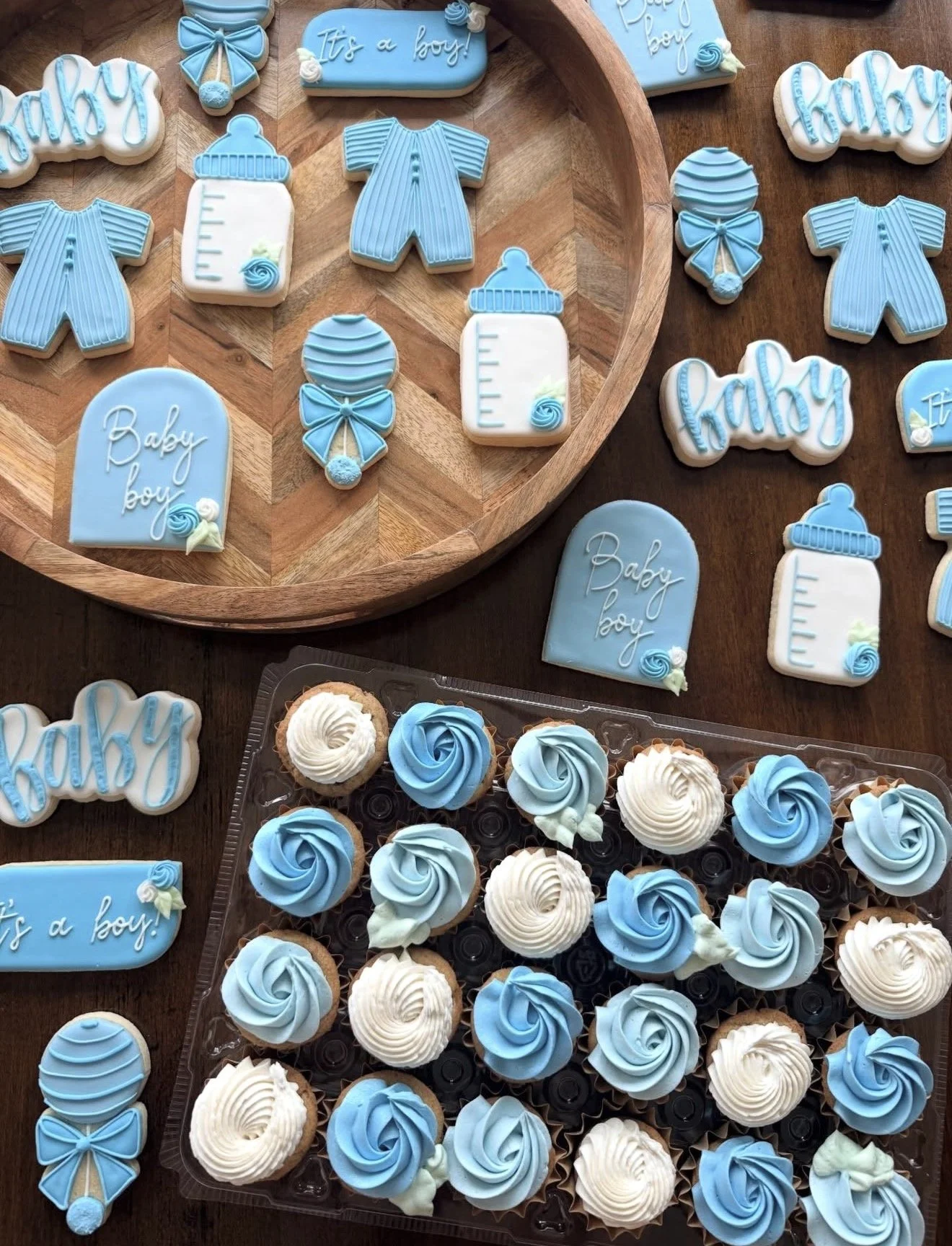 Decorative cookies in blue and white for a baby boy celebration, including ones shaped like baby bottles, onesies, and with inscriptions like 'It's a boy!' and 'Baby boy,' with cupcakes decorated in matching blue and white frosting.