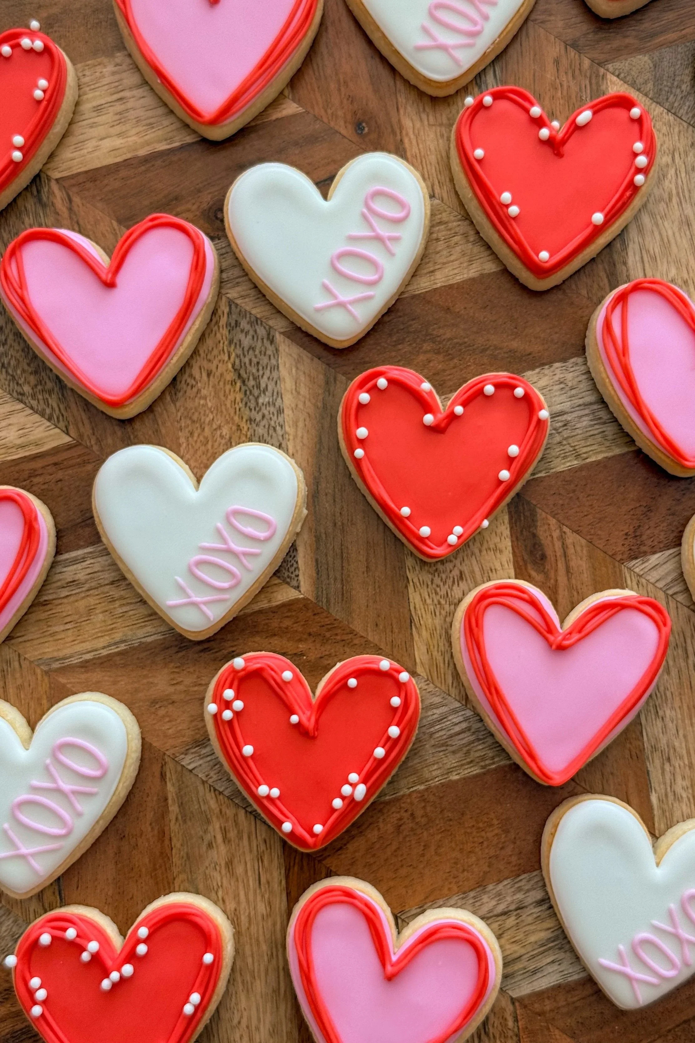 Heart-shaped cookies decorated with pink, red, and white icing, some with the word "XOXO" written in pink, laid out on a wooden surface.