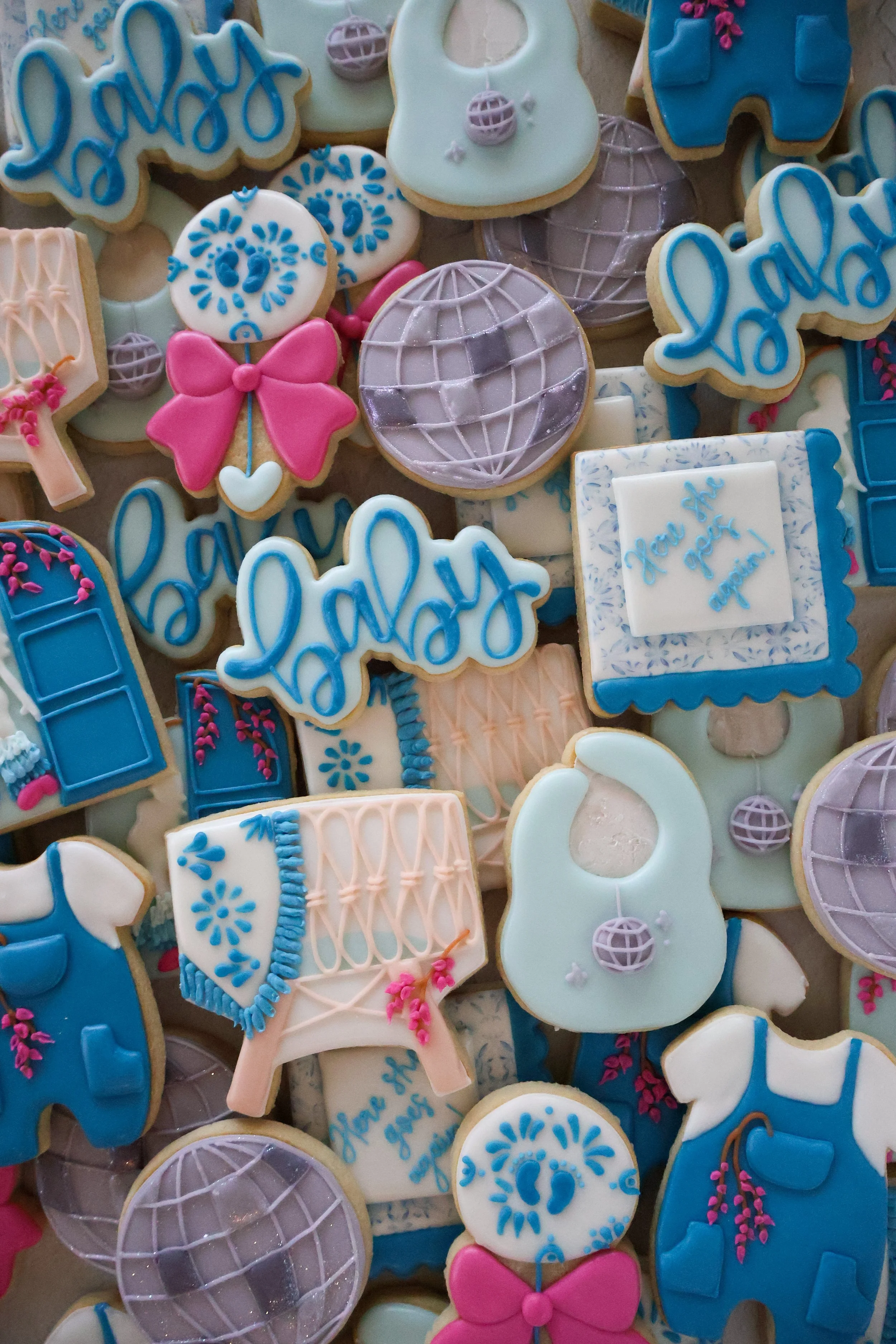 Assorted decorated cookies with blue, pink, and white icing. The cookies have various shapes, including butterflies, globes, and phrases like 'happy' and 'you' with decorative designs.