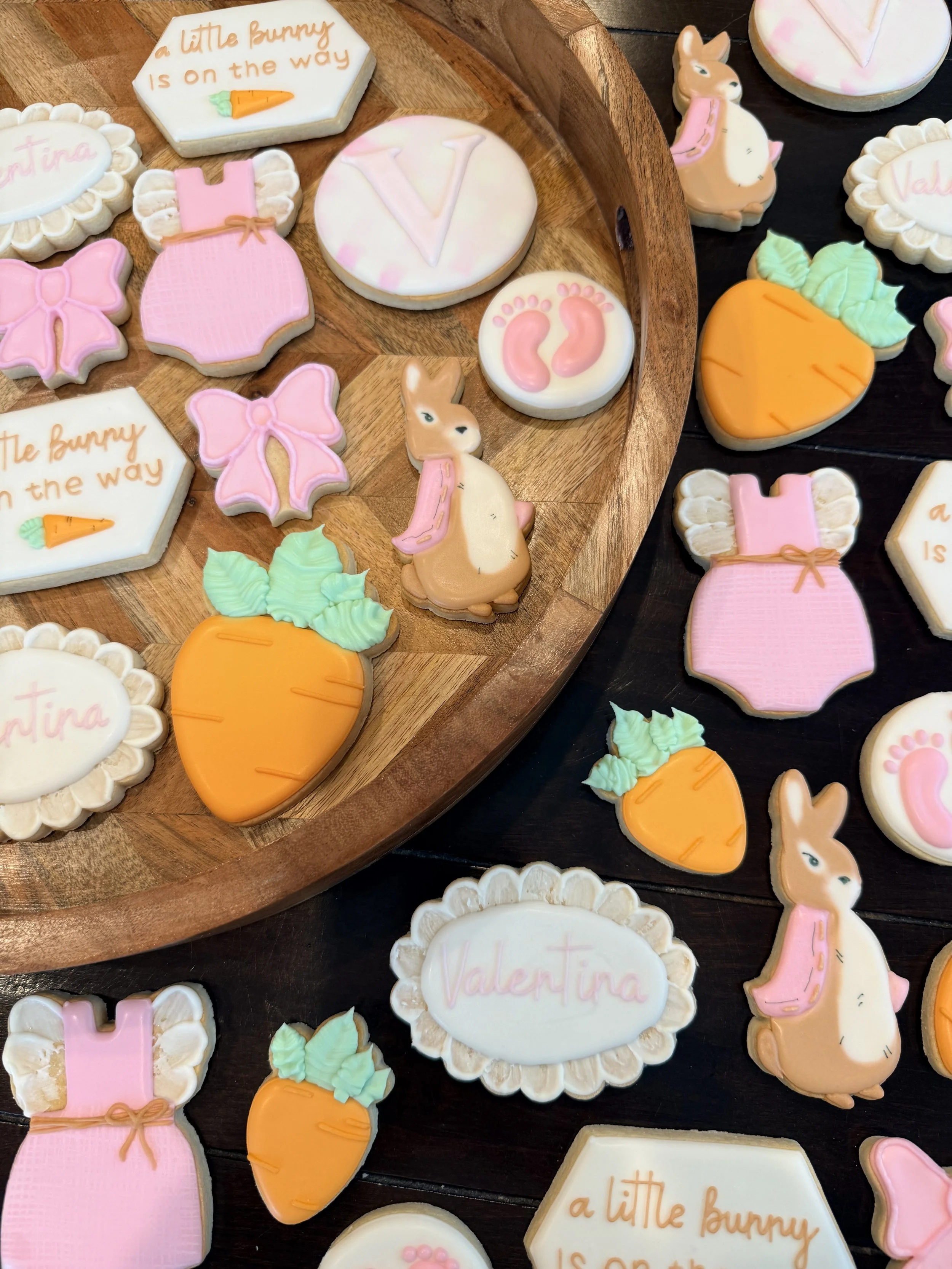 Decorative cookies in pastel colors with Easter and spring theme. Some cookies have messages like 'a little bunny is on the way' with a carrot drawing, and others have names such as 'Valentina'. There are cookies shaped as bunnies, carrots, and bows,