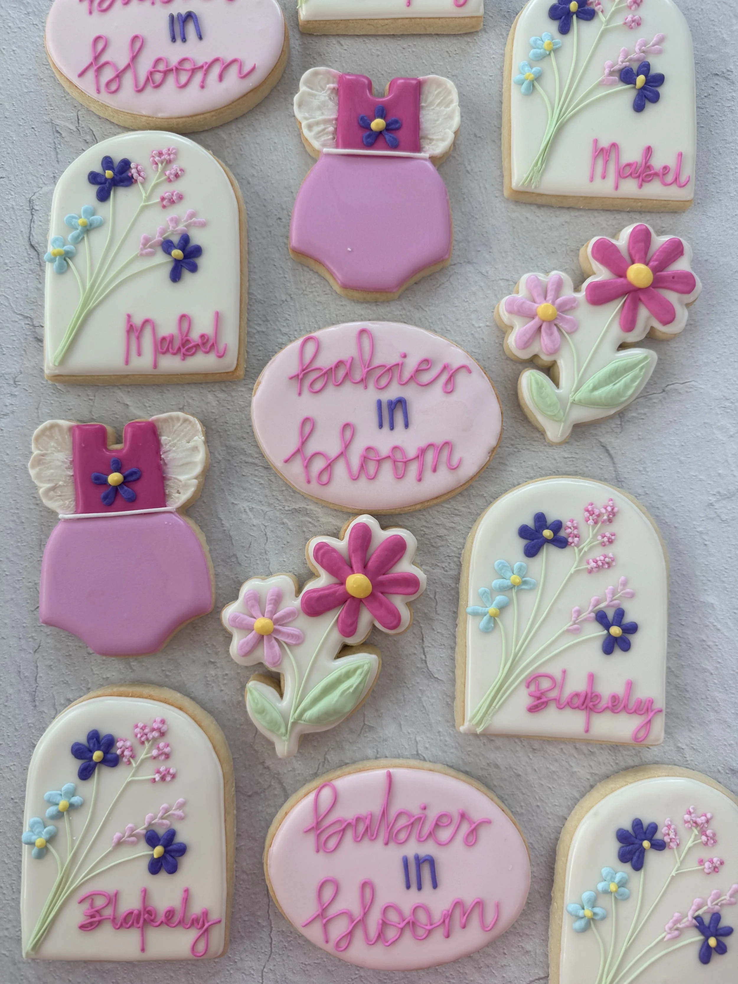 Decorated cookies with pastel pink, purple, and white icing, featuring floral designs, baby bottle shapes, and messages like 'babies in bloom' and 'blessed' in pink and purple icing.