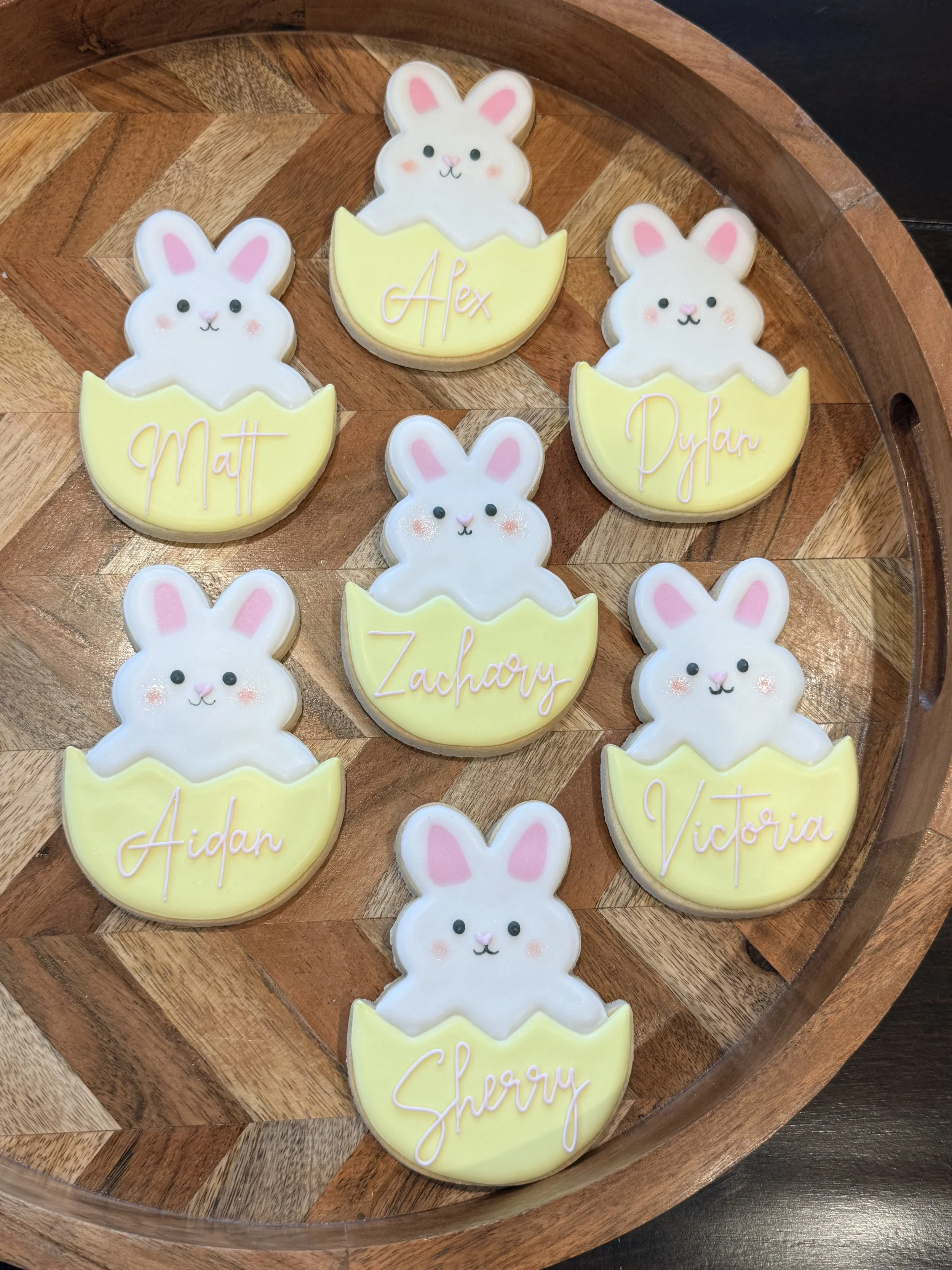 Cookies shaped like bunnies with yellow eggs, decorated with pastel pink, white, and black icing, each cookie has a name written in pink icing, including Matt, Alex, Dylan, Aidan, Victoria, and Sherry, placed on a round wooden tray.