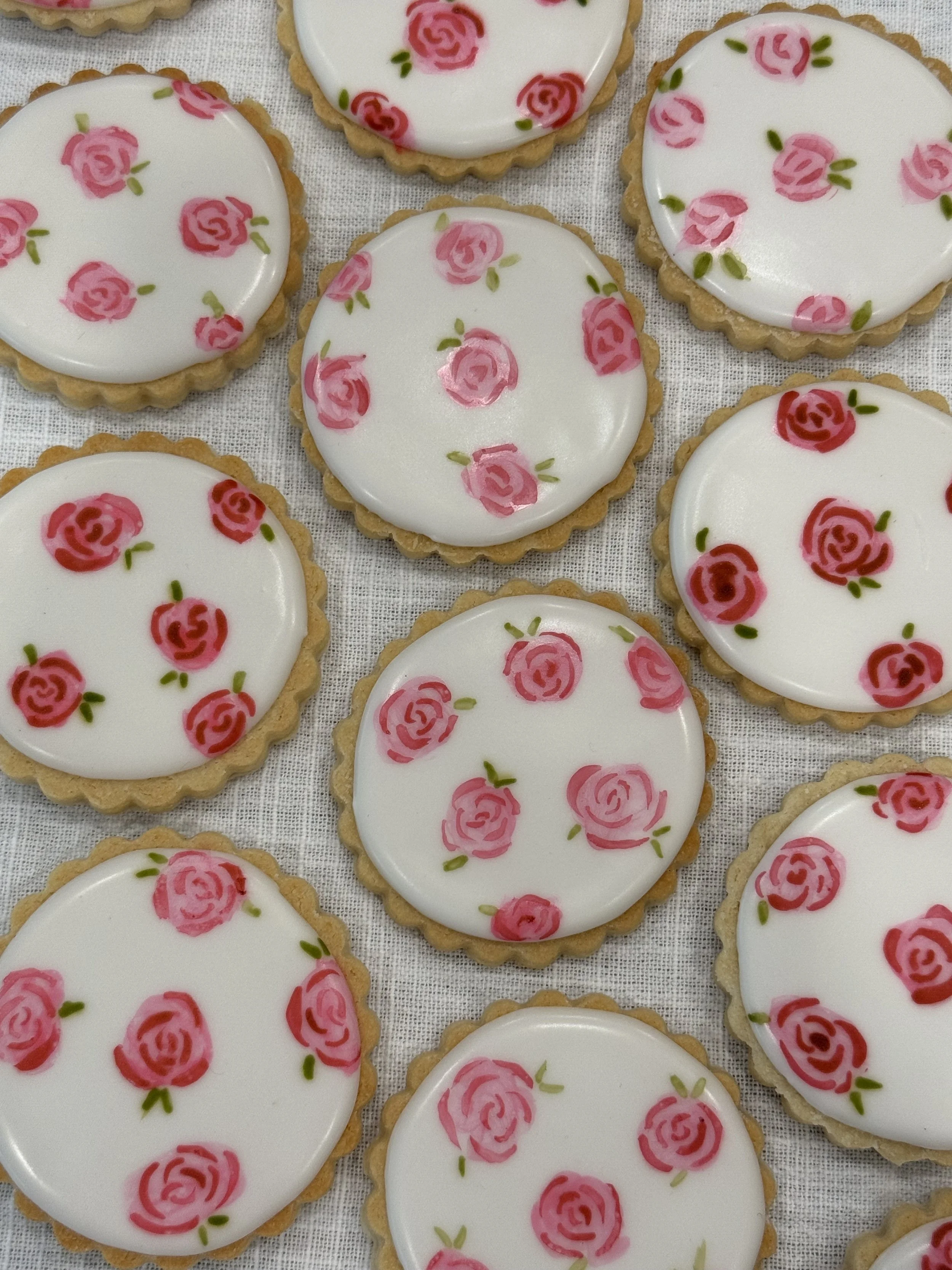 A collection of oval-shaped cookies with a scalloped edge, decorated with white icing and pink rose patterns with green leaves.
