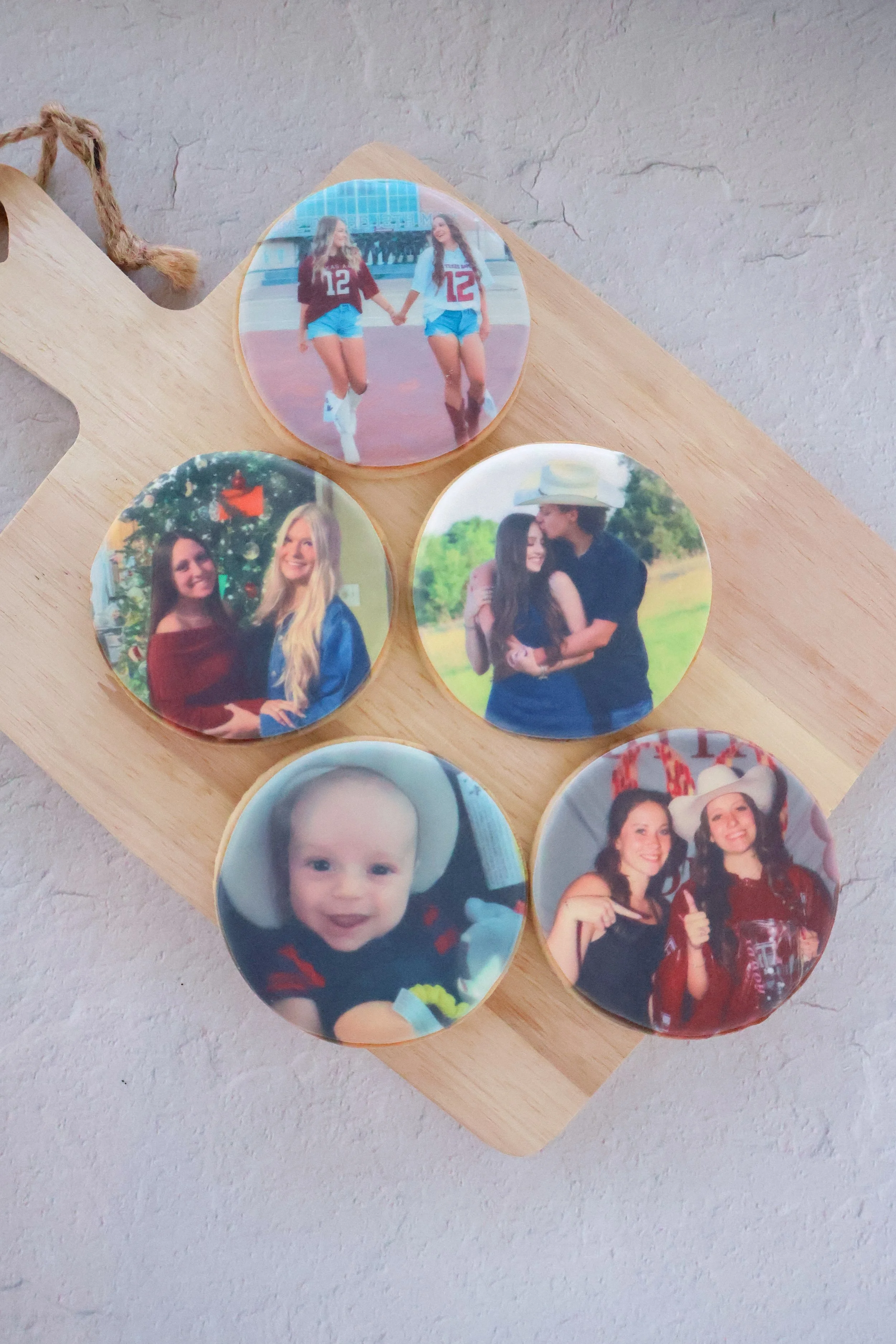 A wooden board holding various round photo magnets featuring people, including two women holding hands at a track field, two women smiling at a decorated Christmas tree, two women hugging outdoors, a smiling baby, and two women dressed up at a party.