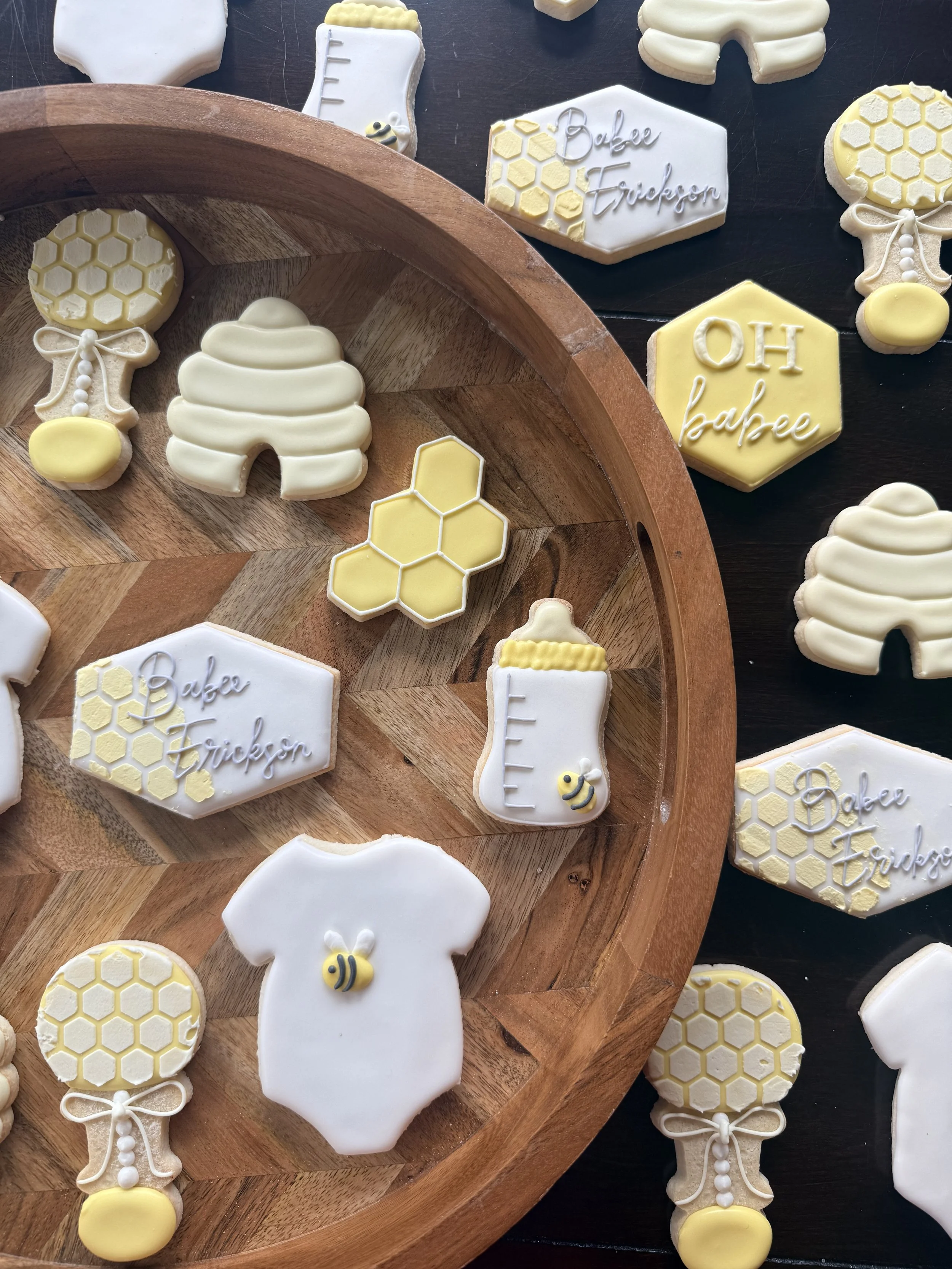 Cookies decorated with honeycomb patterns, bees, and honey jars, arranged on a wooden tray and black surface.