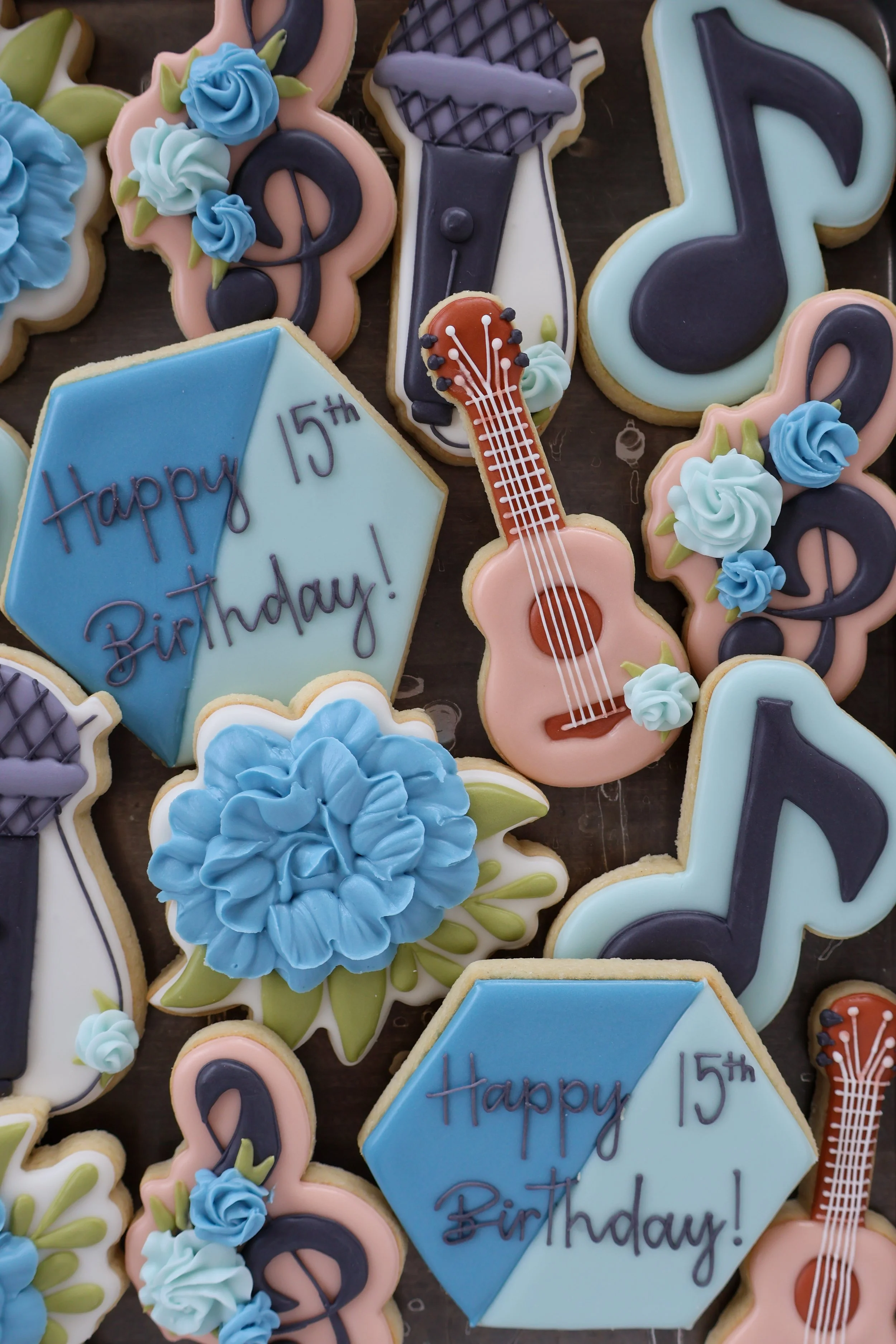 Decorative birthday cookies with musical and floral themes, including a cake with the message "Happy 15th Birthday!" written on a blue diamond-shaped cookie, cookies shaped like guitars and musical notes, and floral designs with blue and white icing.