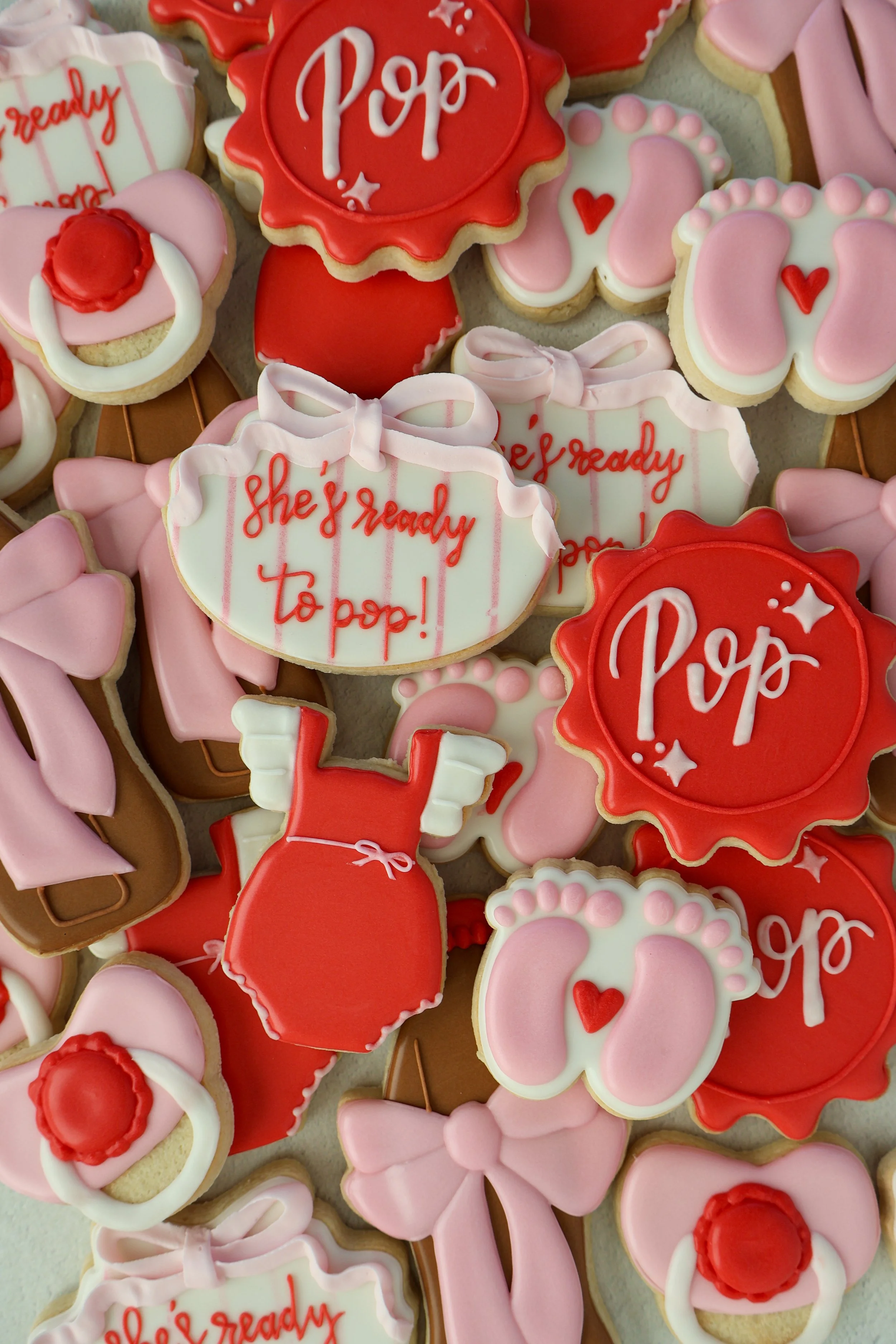 Decorated cookies with various pink, red, and white icing, including hearts, bows, and messages such as 'she's ready to pop!' and 'Pop'.