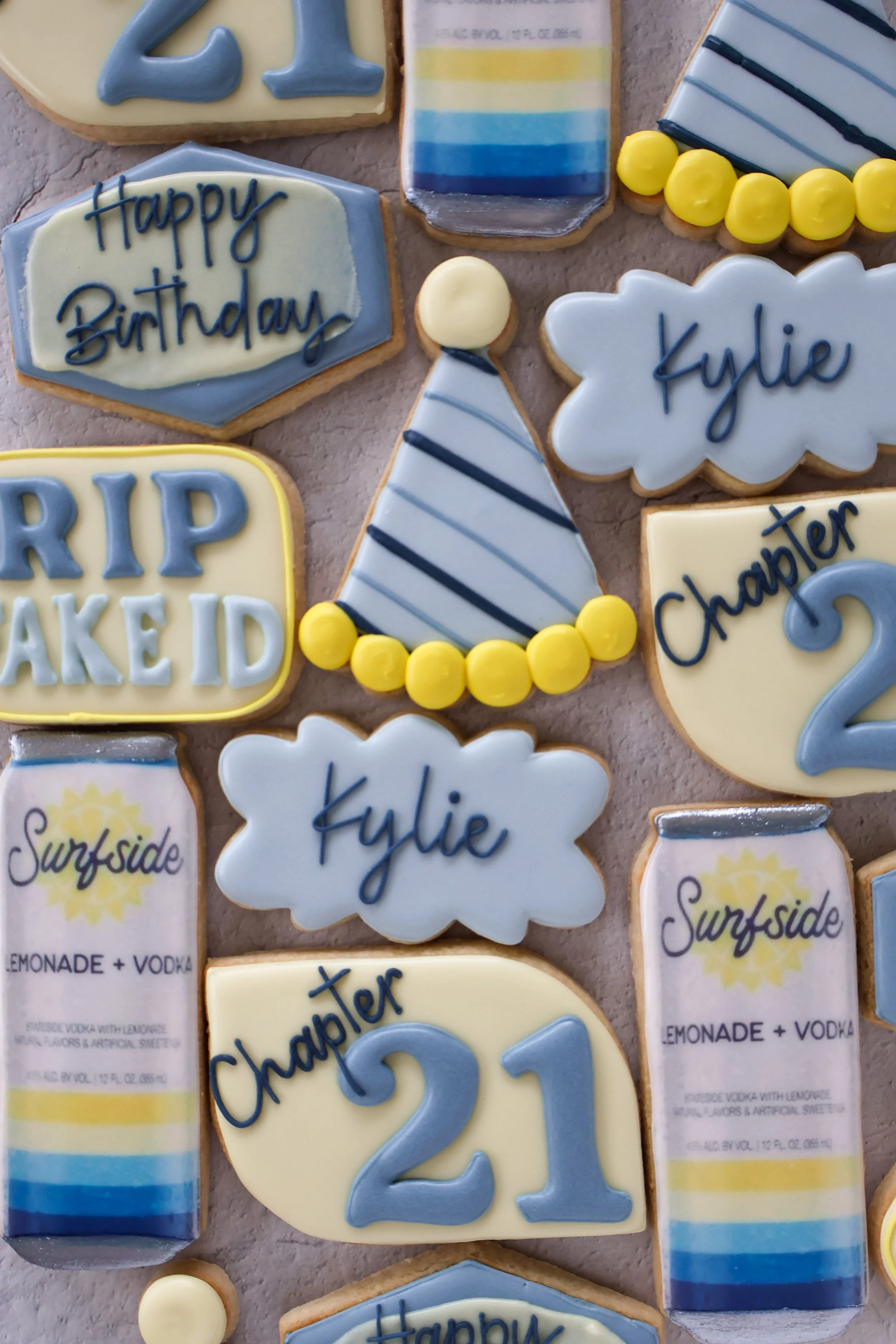 Decorative birthday cookies with various shapes and messages in blue, yellow, and white icing. Some cookies say 'Happy Birthday,' 'Kylie,' 'Chaper 2,' and 'RIP AKEID,' along with colorful birthday-themed designs like a party hat, a rainbow, a cloud, 