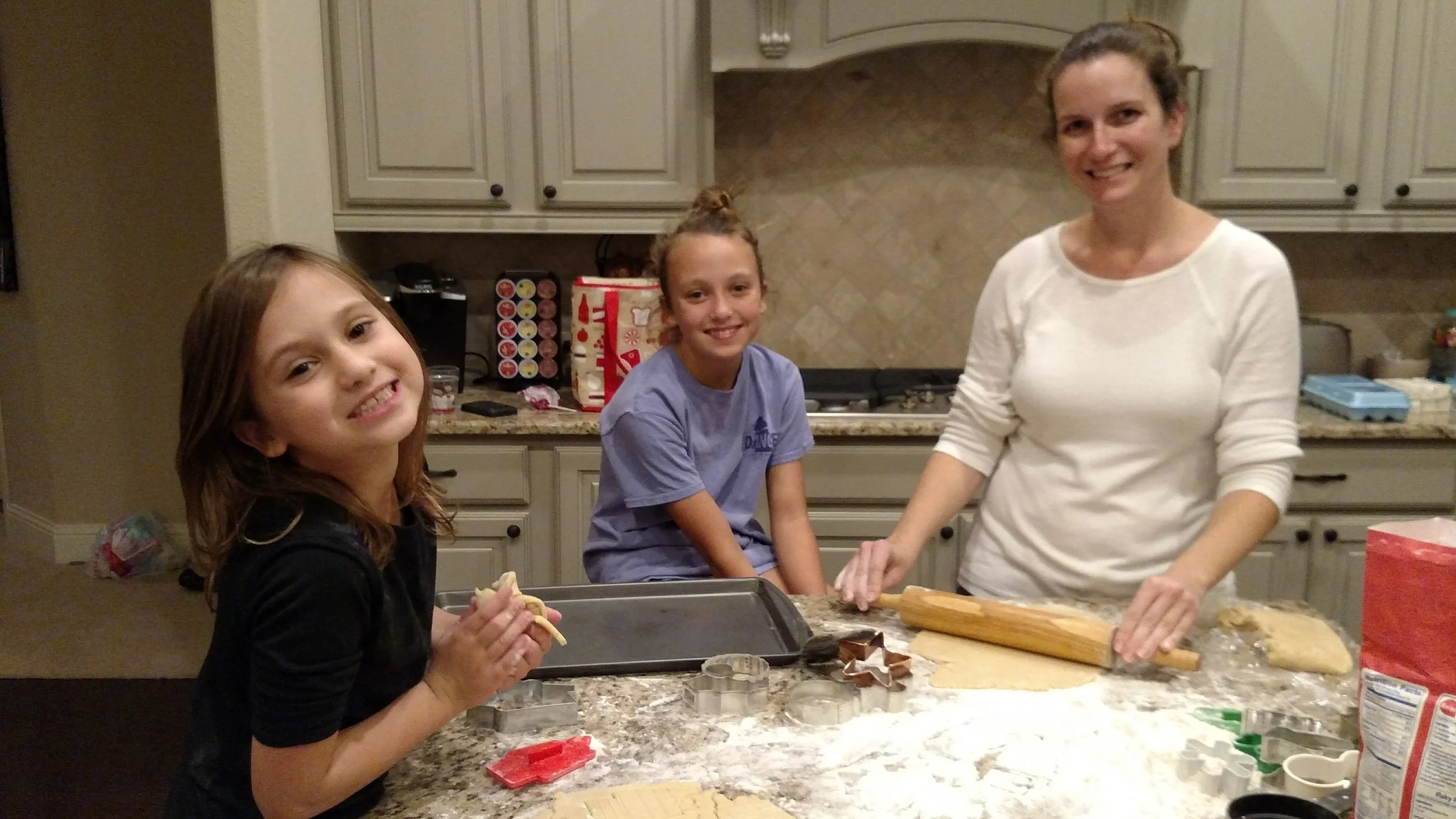 Three females in a kitchen preparing holiday cookies, with holiday-themed items and baking supplies on the counter.