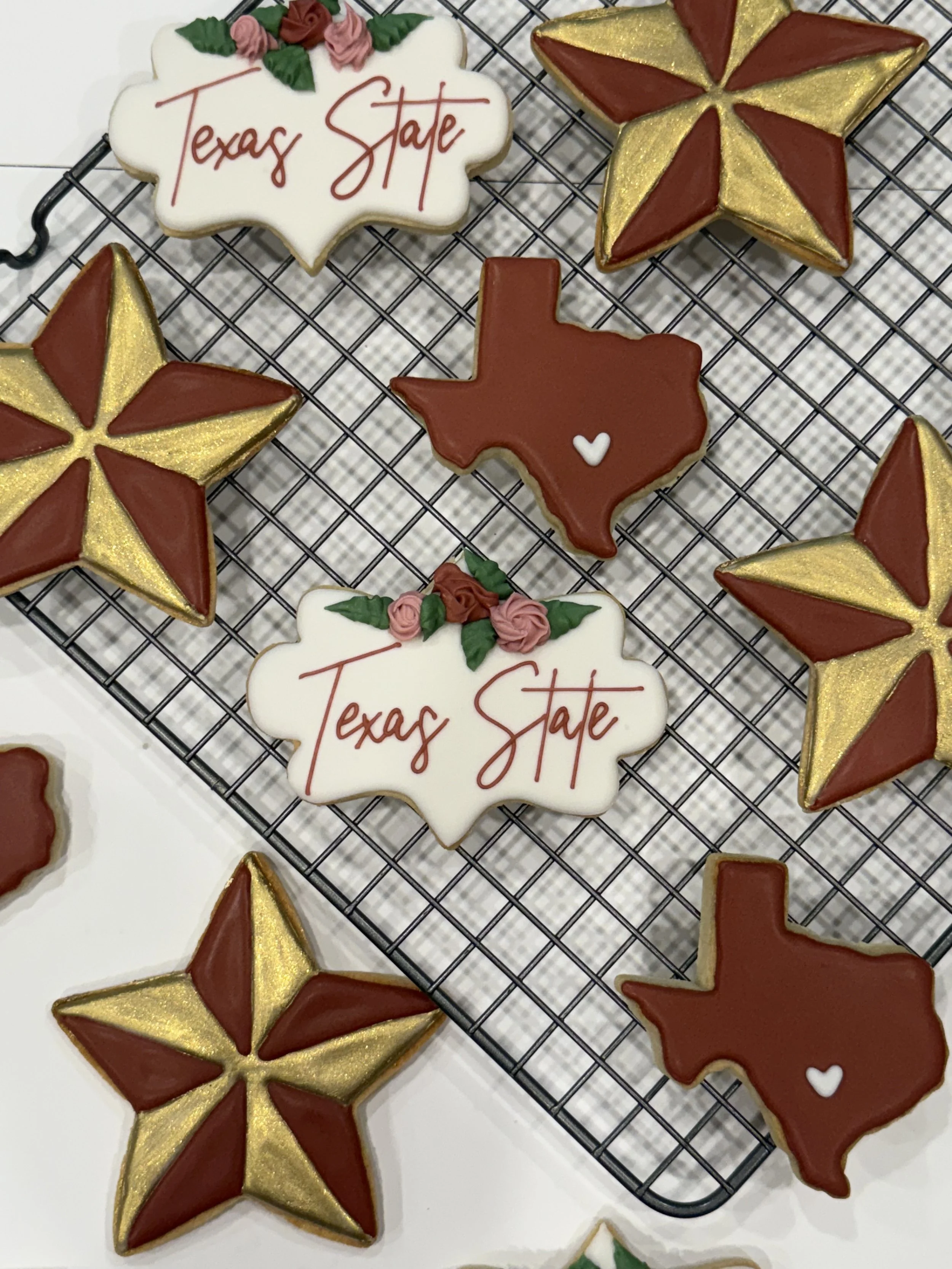 Decorated cookies on a cooling rack, including a Texas-shaped cookie with a small white heart, star-shaped cookies with red and gold icing, and a cloud-shaped cookie with pink roses and green leaves, with the words 'Texas State' written in red.