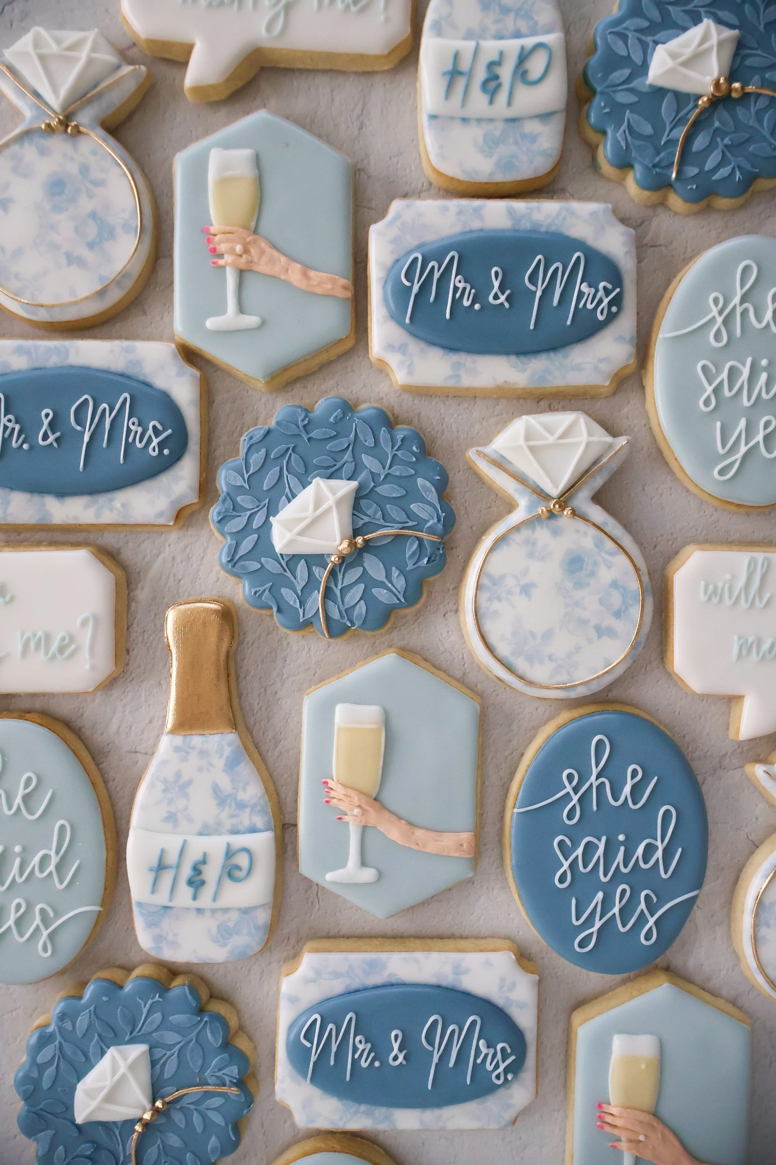 Collection of wedding-themed decorated cookies in blue, white, and gold. Some cookies have inscriptions like 'Mr. & Mrs.' and 'she said yes,' while others feature designs of champagne glasses, engagement rings, and floral patterns.