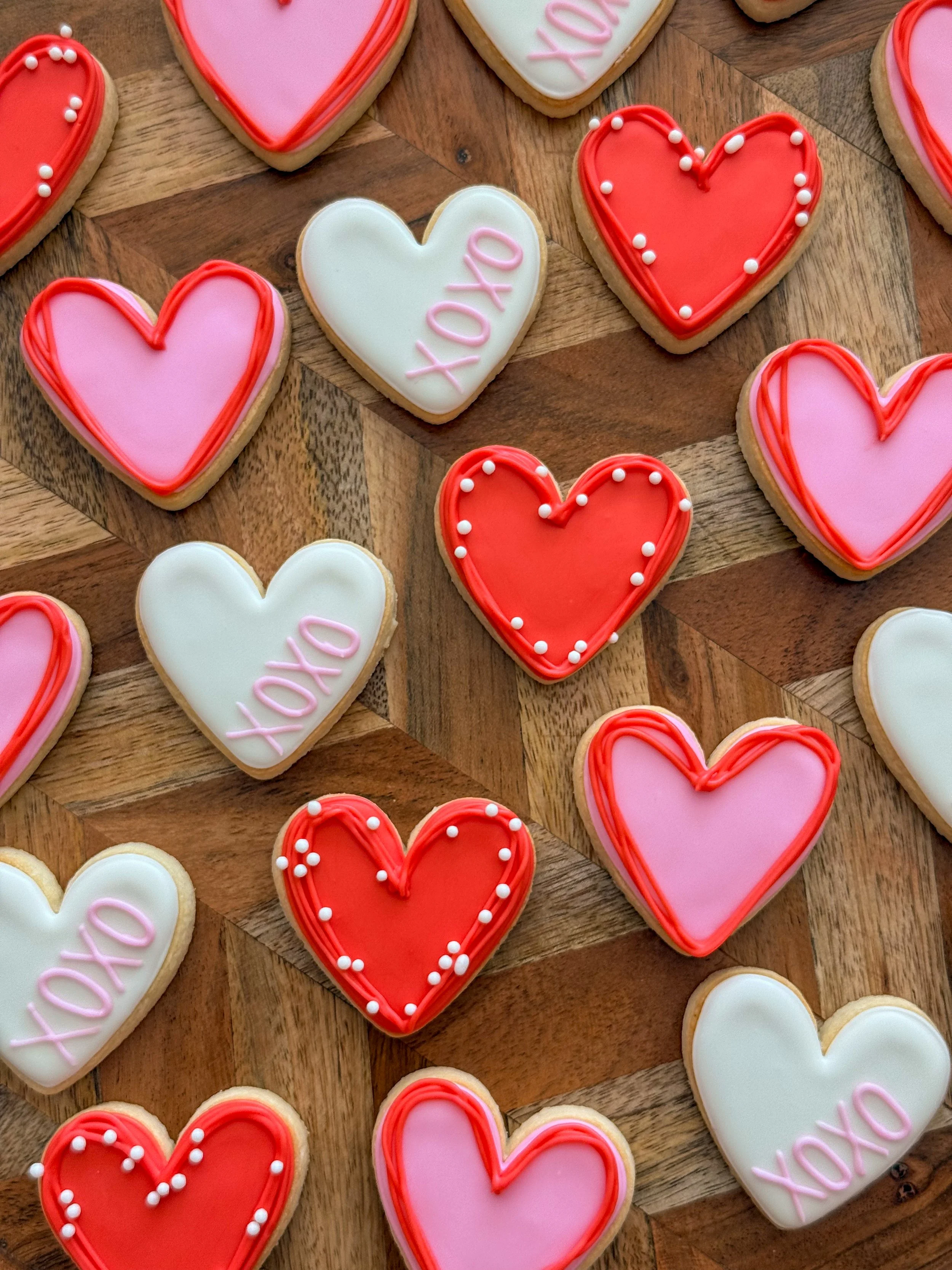 Heart-shaped cookies decorated with pink, red, and white icing, some with "XOXO" written on them, on a wooden surface.